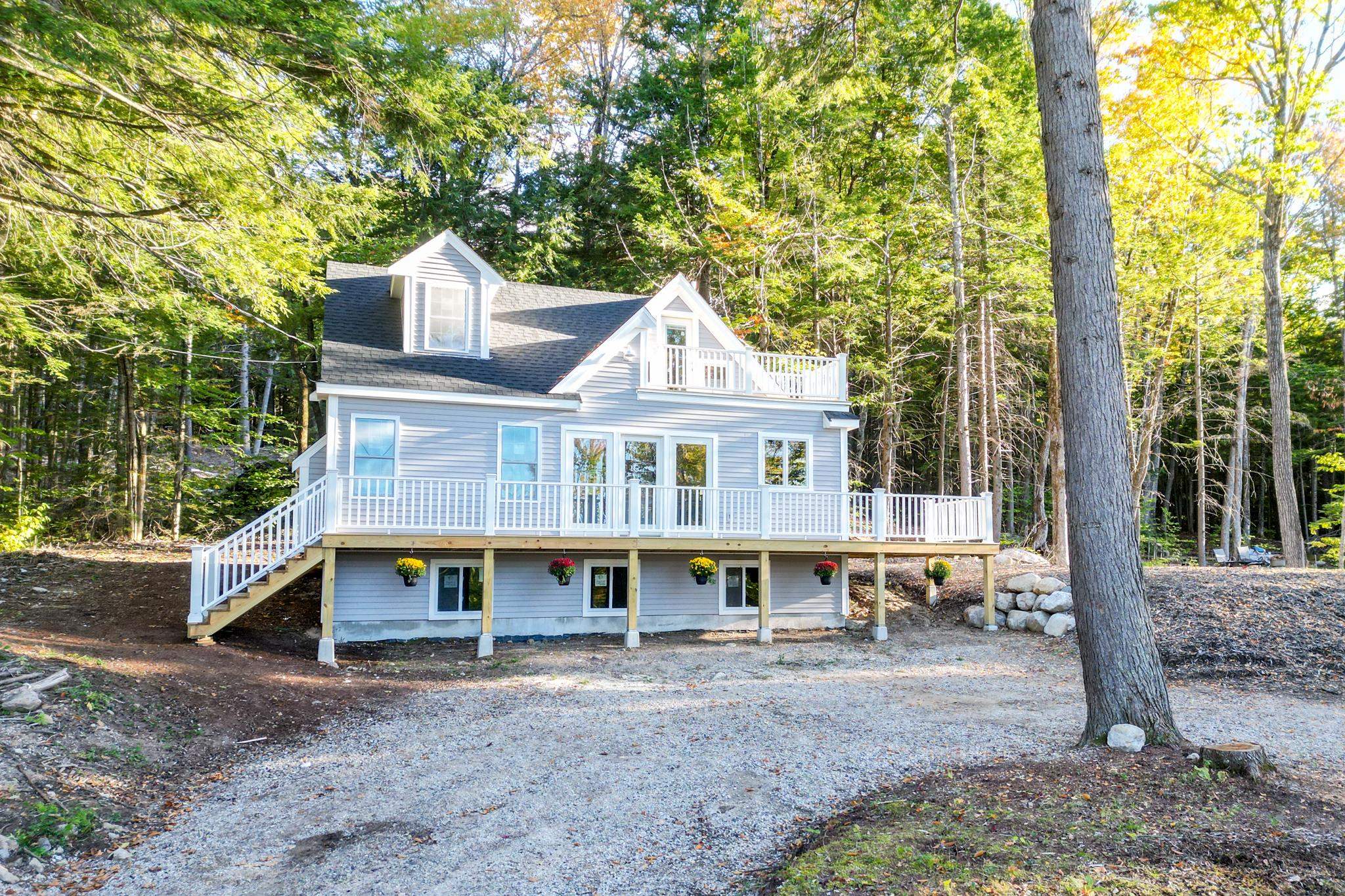 Light gray house with a raised wraparound deck and upper balcony, surrounded by trees.
