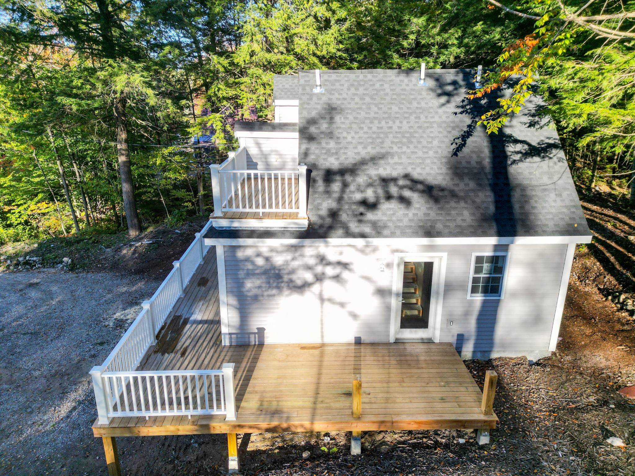 Rear of a light-colored house with a large wood deck, upper balcony, and wooded surroundings.