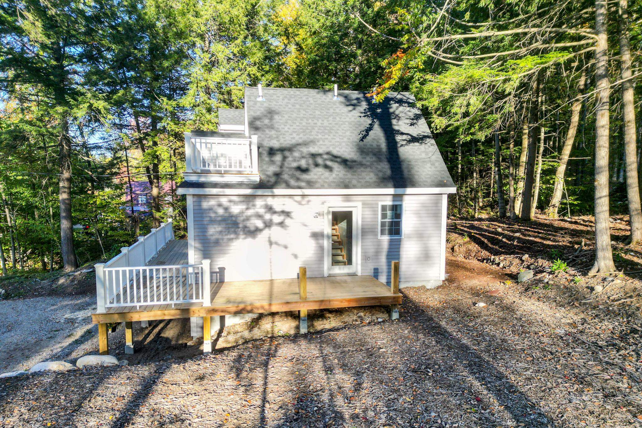 Gray house with a steep roof and raised deck among trees on a leaf-covered lot.