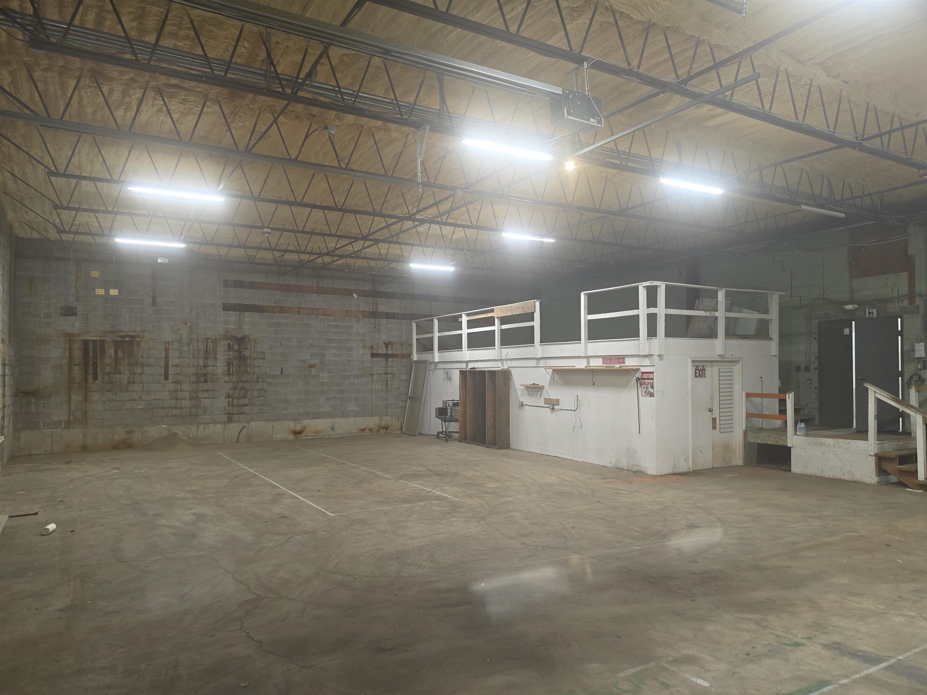 Large vacant warehouse interior with exposed roof trusses, concrete floor, and a raised office platform.