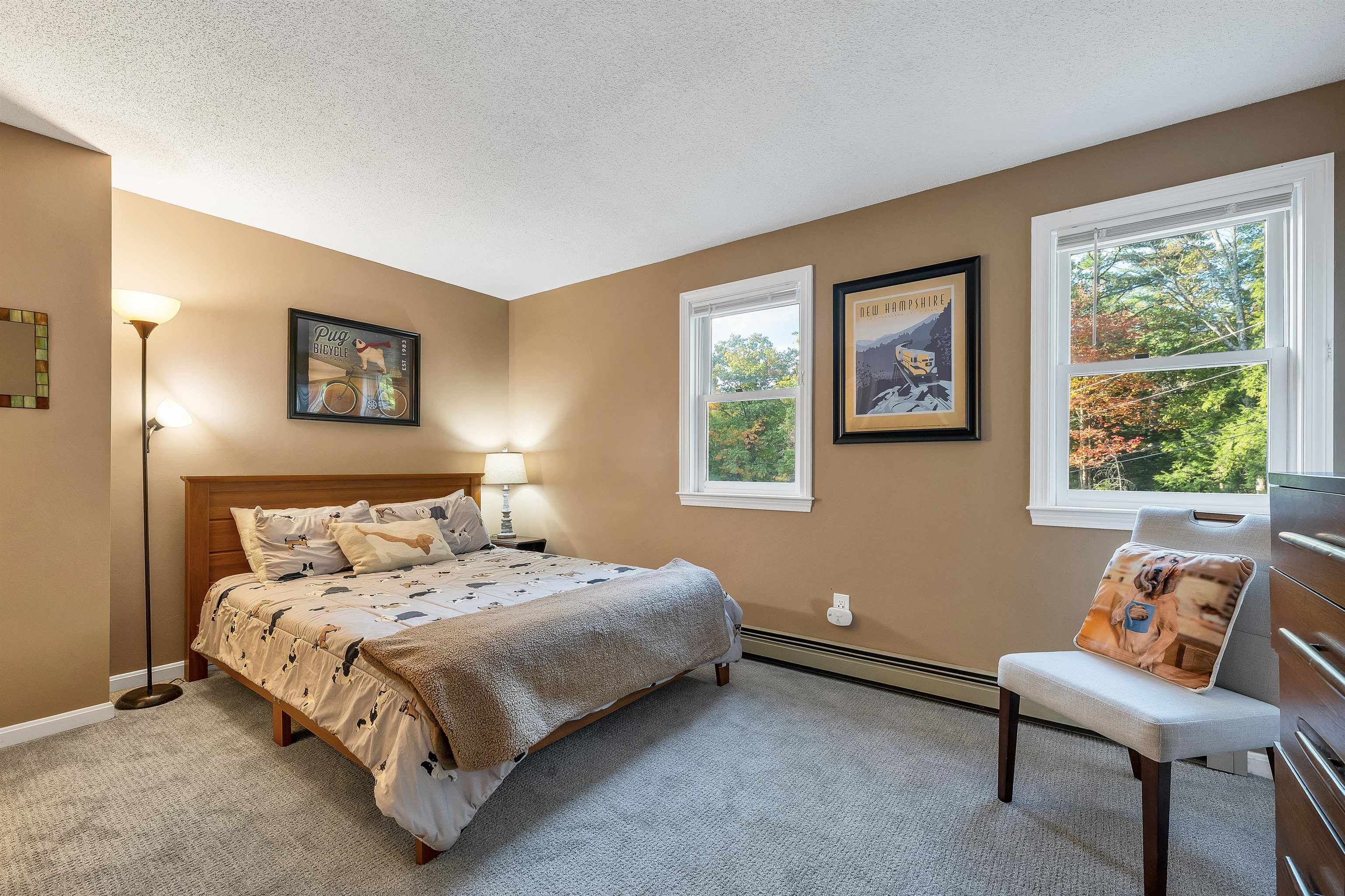 Bedroom with dog-print bedding, a cushioned chair, and windows overlooking autumn foliage.