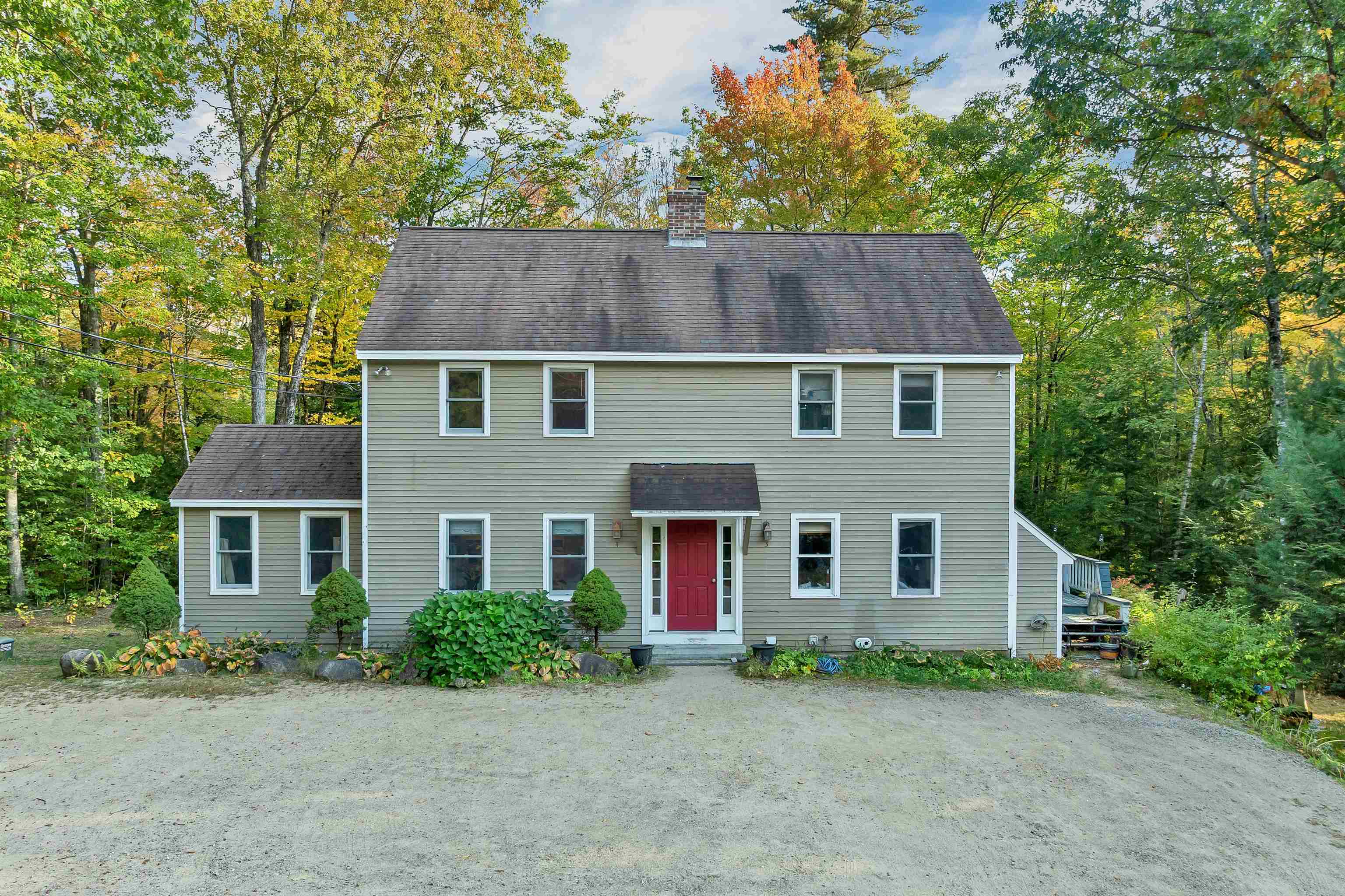 Two-story gray-green house with a red front door, gravel driveway, and autumn trees.