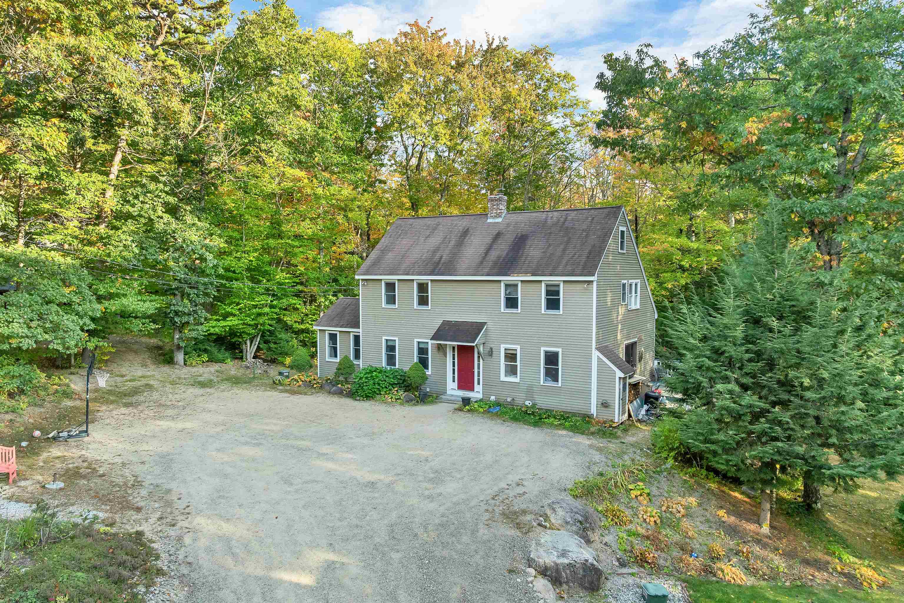 Two-story gray-sided house with a red front door, surrounded by autumn trees and a broad gravel driveway.