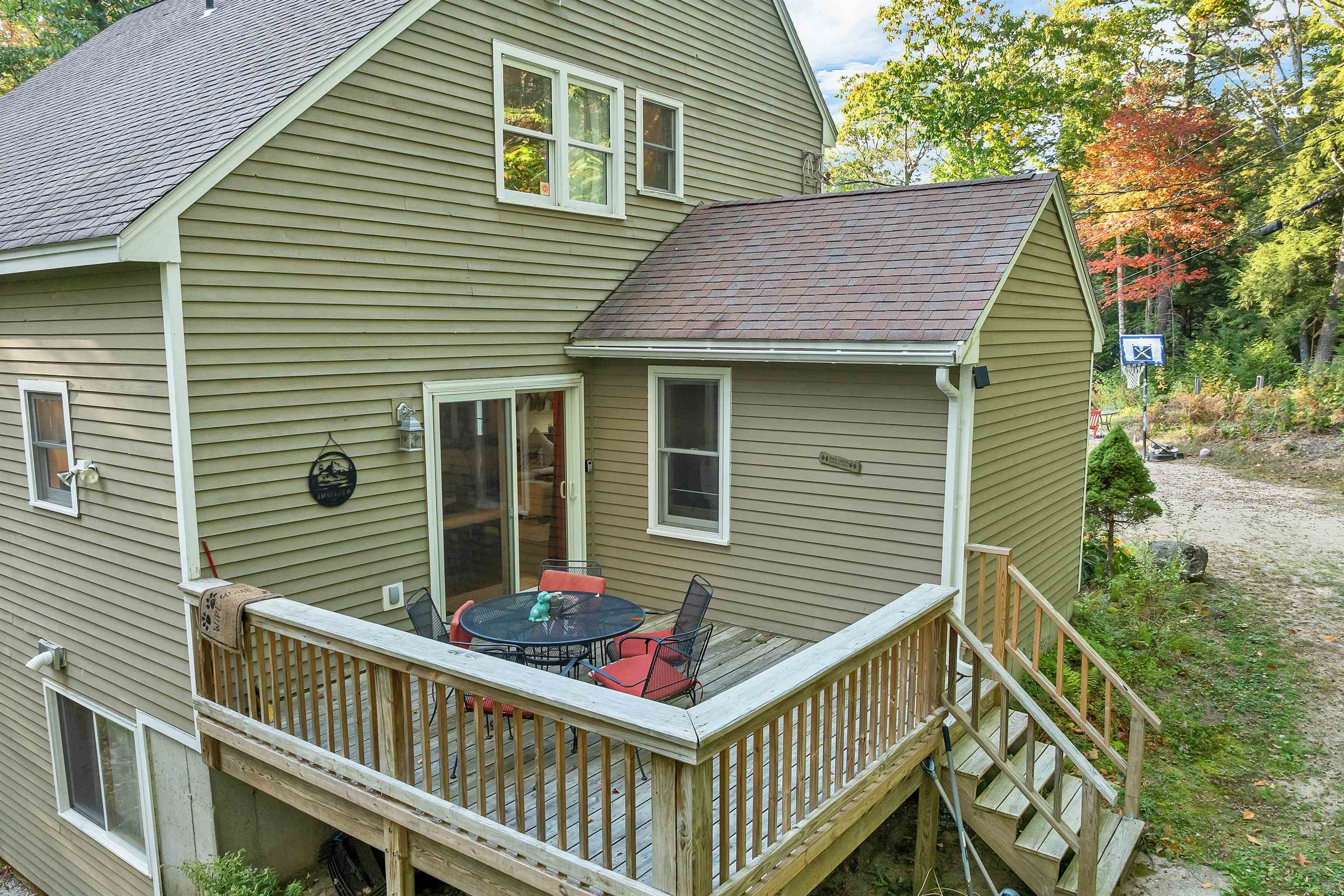 Home’s rear deck with outdoor dining furniture, wooden stairs, and a basketball hoop beside the wooded yard.