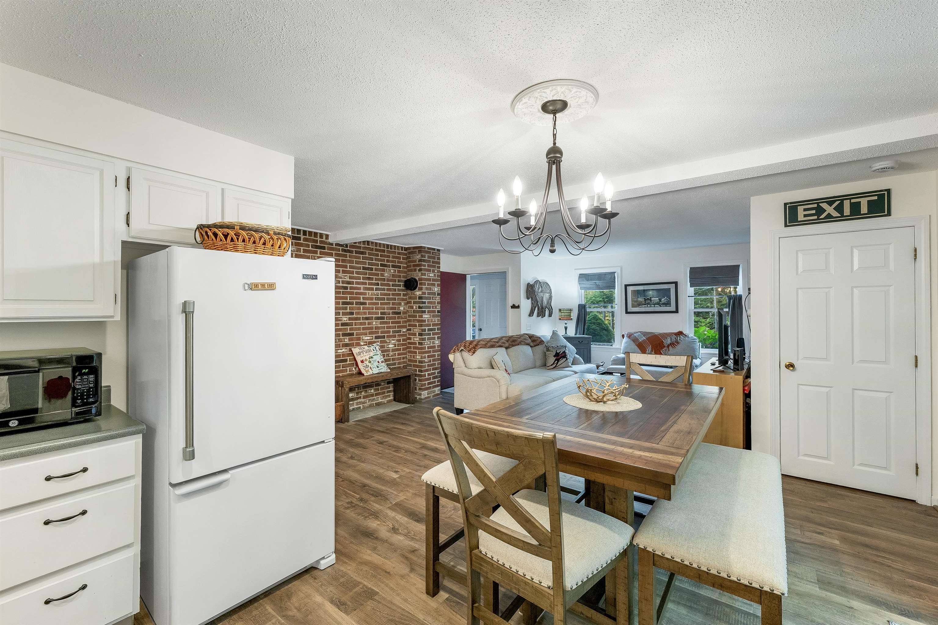 Open-plan dining area with a wood table, beside a kitchen and brick fireplace.