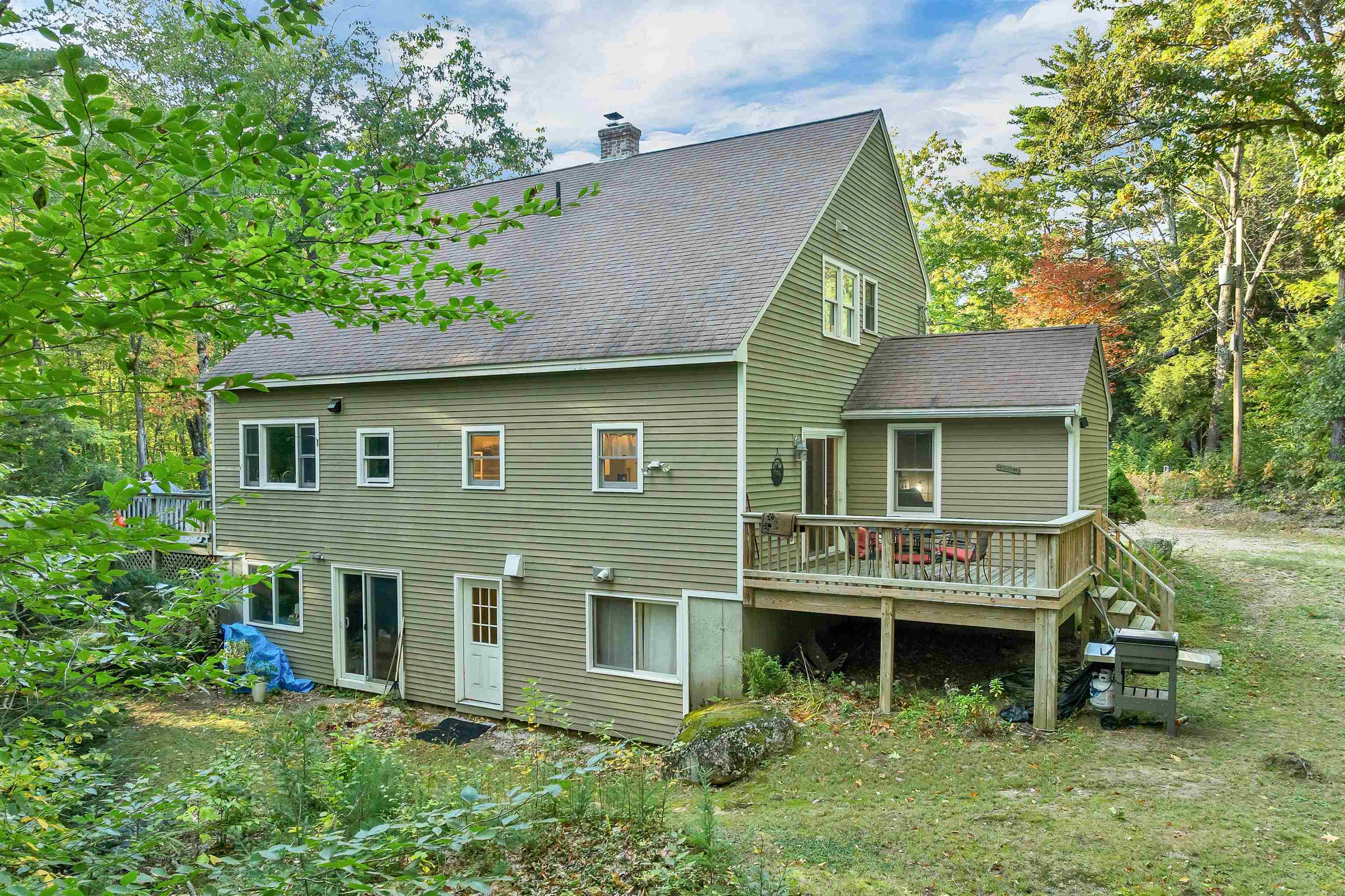 Green-sided house with a raised deck and outdoor seating in a wooded yard.