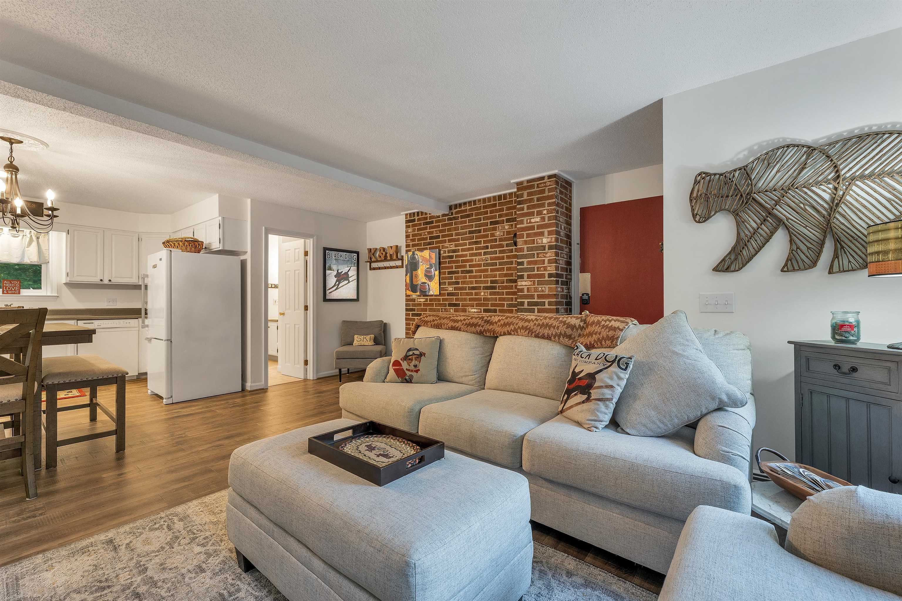 Open-plan living room with a gray sectional, exposed brick wall, and adjoining white kitchen.