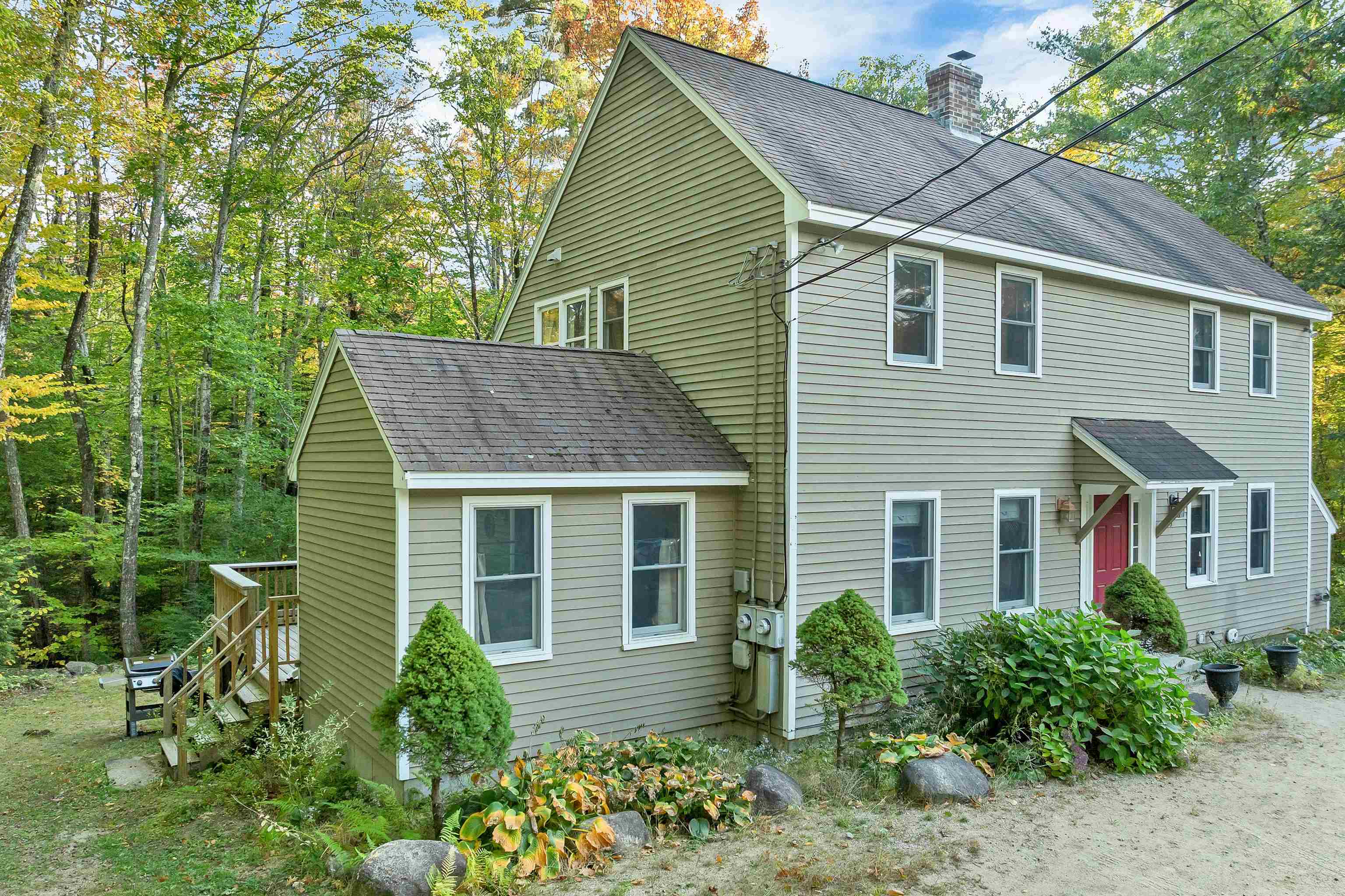 Two-story gray-green house with a red door, covered entry, and wooded autumn surroundings.