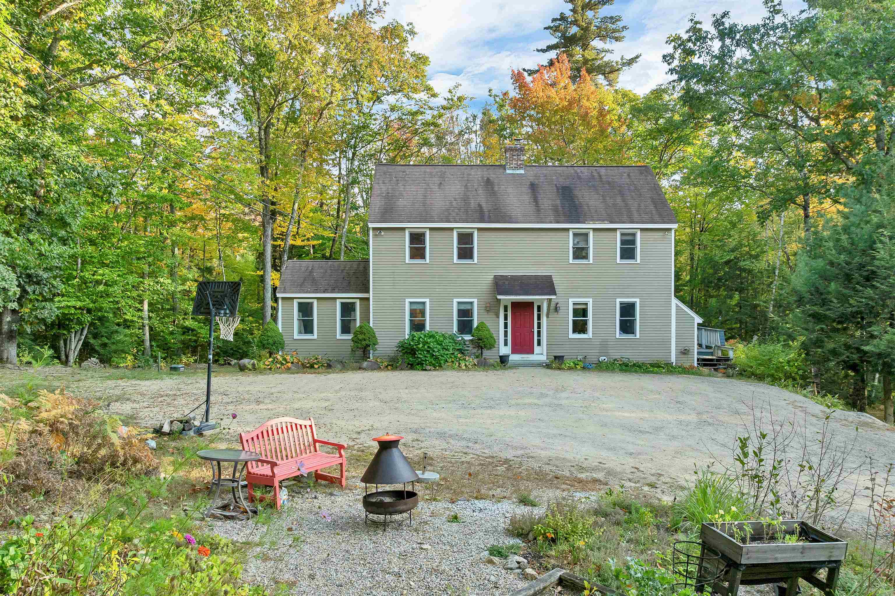 Two-story home with a red front door, gravel yard, basketball hoop, and fire pit among autumn trees.