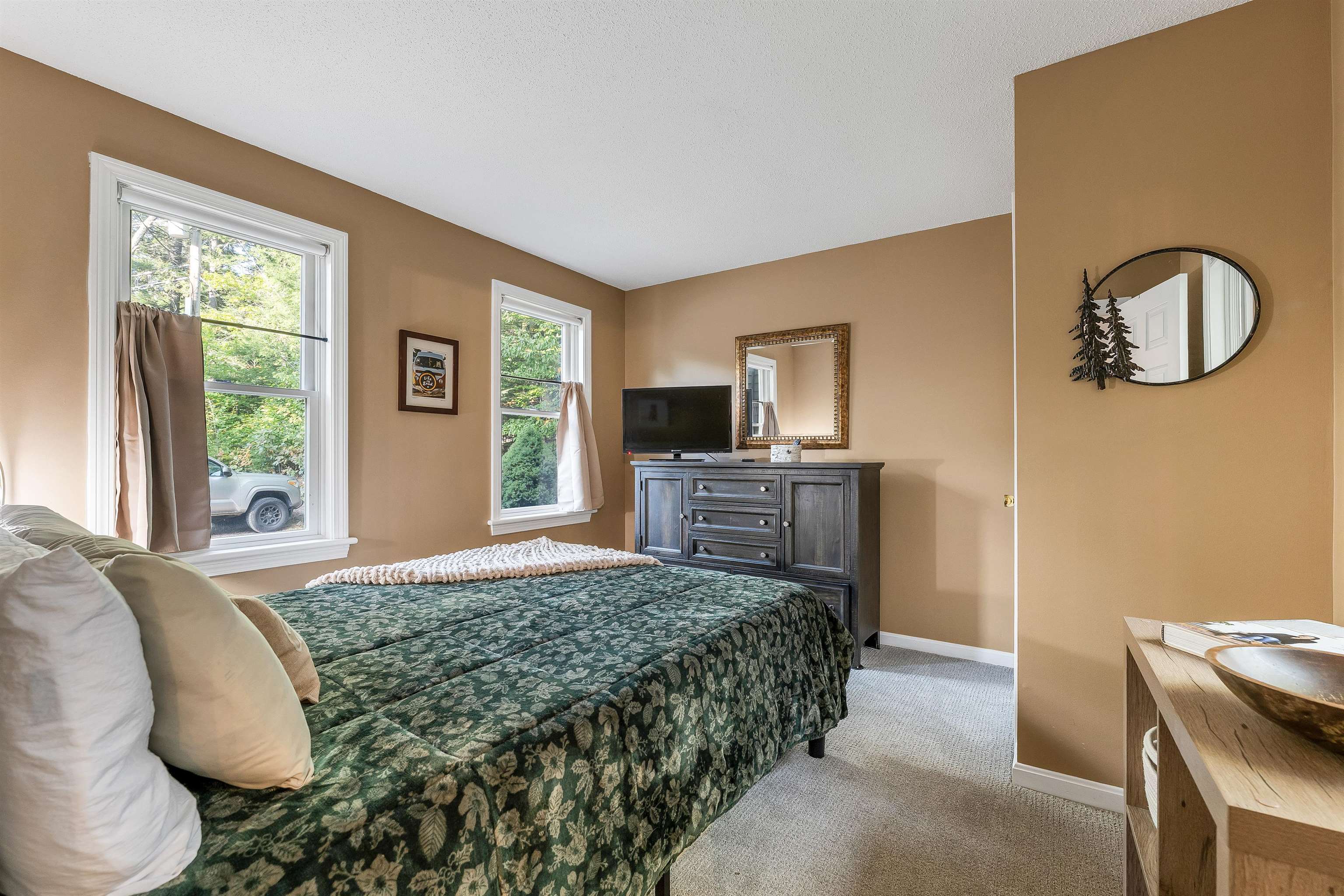 Bedroom with a green floral bedspread, tan walls, and two bright windows.