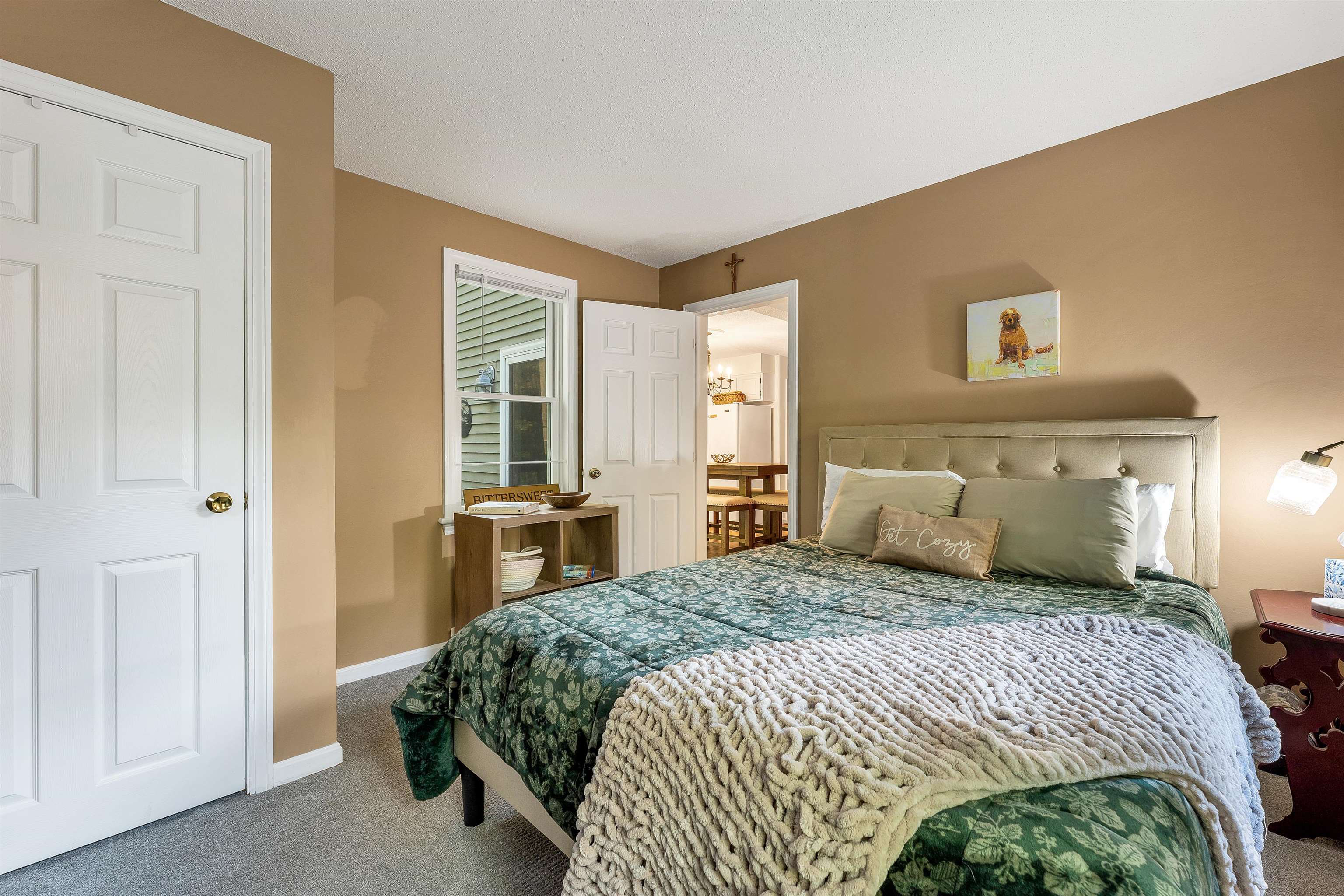 Furnished bedroom with a green floral bedspread, cream throw, and open doorway to the dining area.