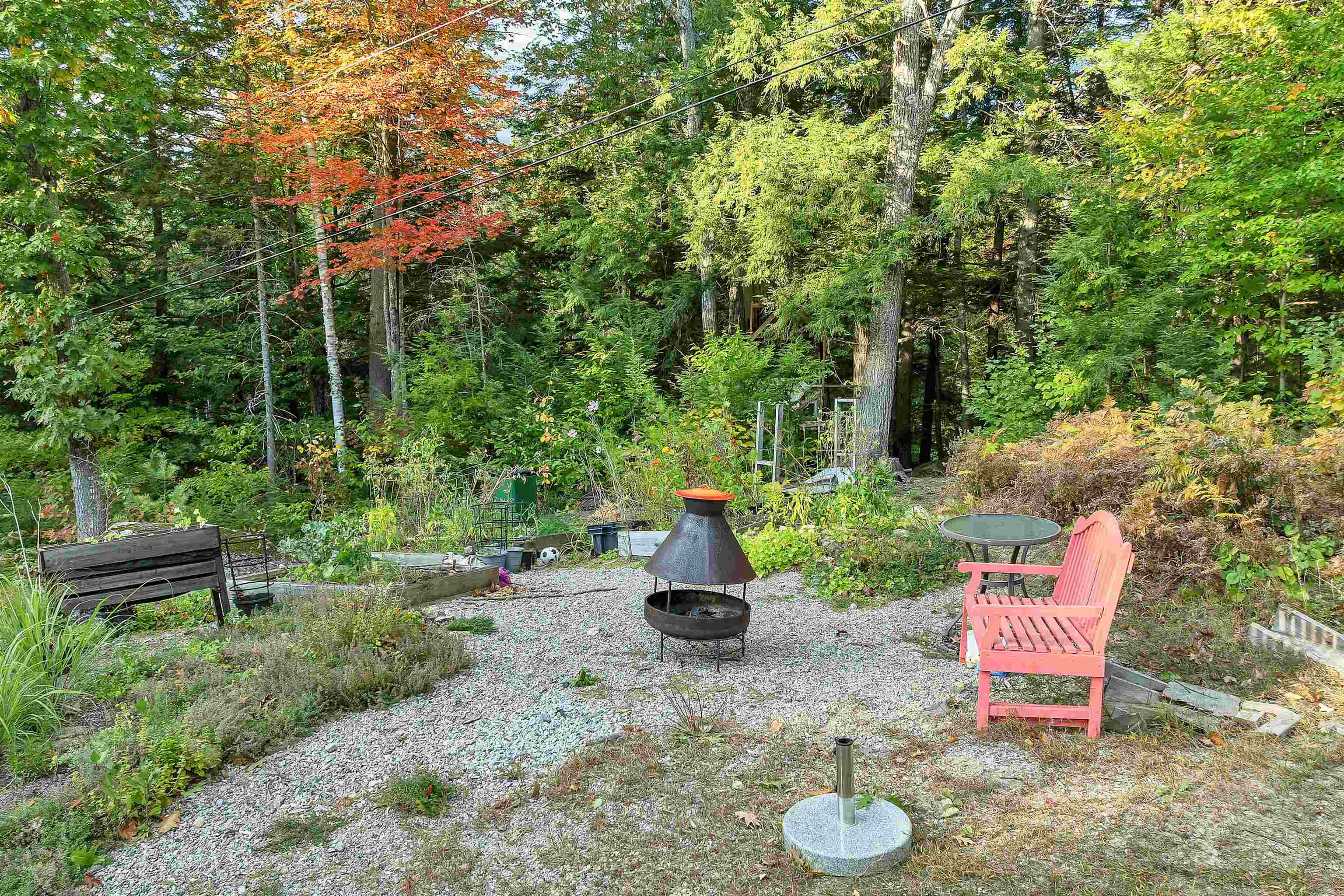 Gravel patio with a metal fire pit and coral Adirondack chair beside wooded garden beds.