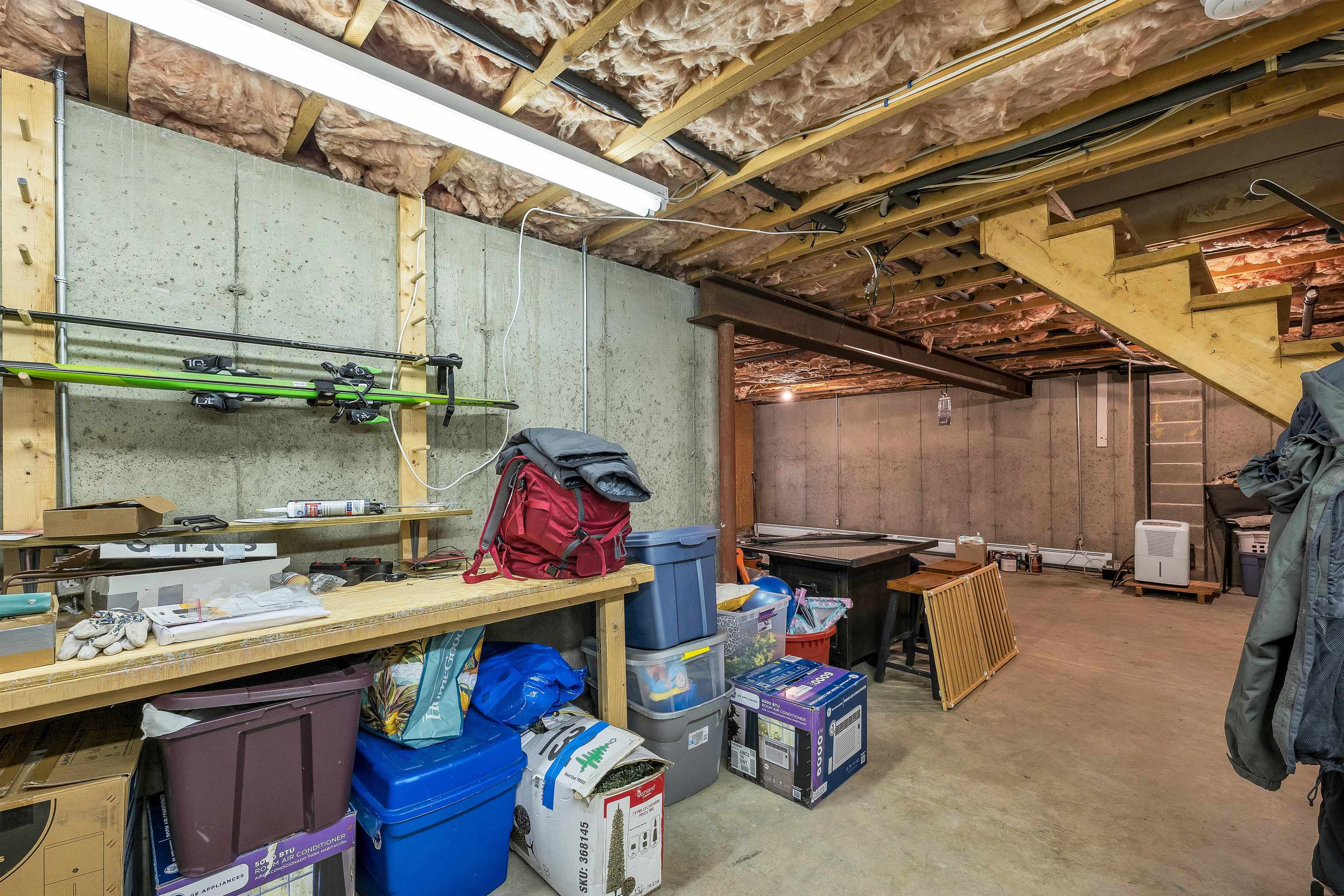 Unfinished basement with exposed insulation, wooden stairs, a workbench, skis, and storage bins.