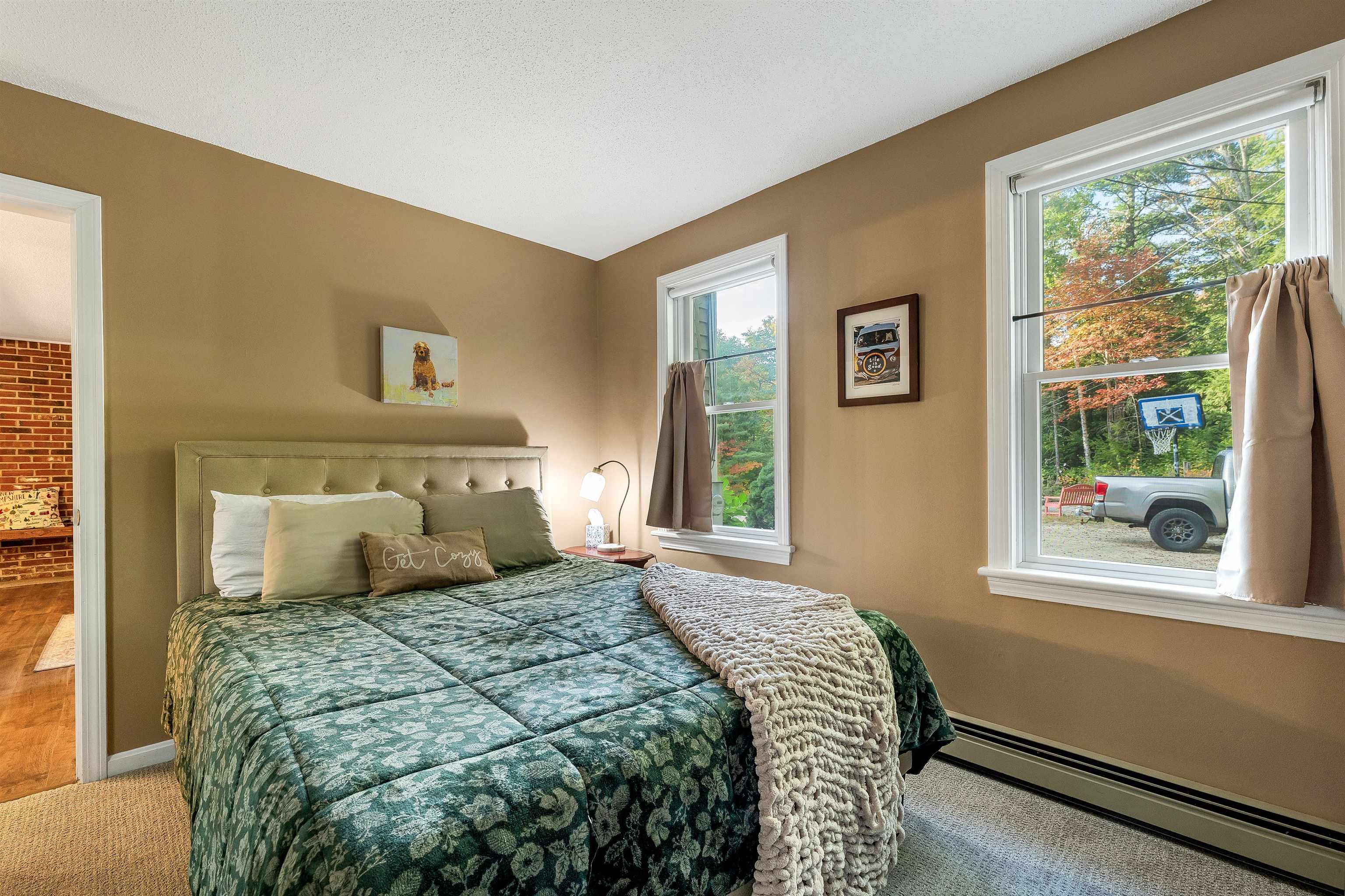 Furnished bedroom with a green floral bedspread and windows overlooking autumn trees.