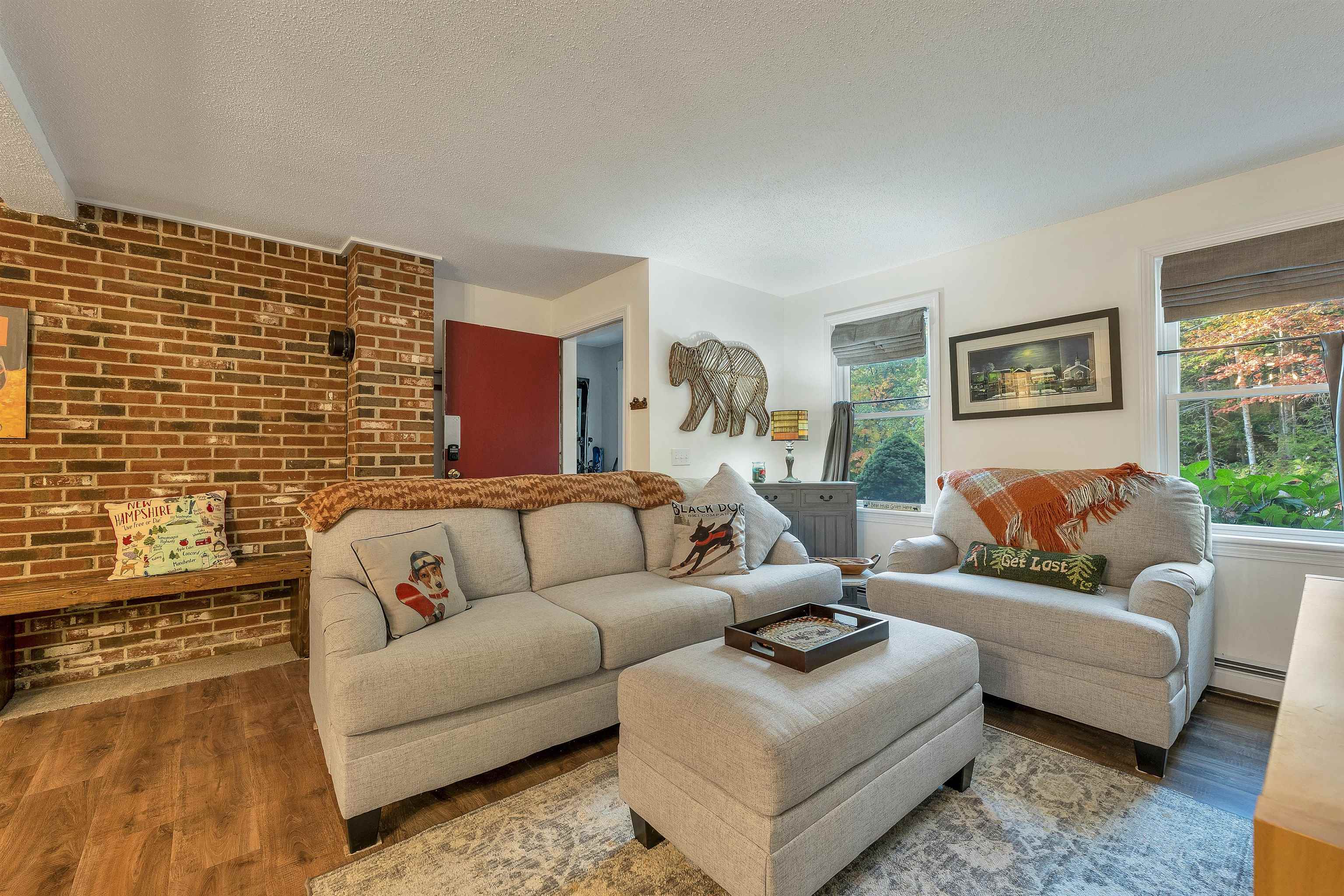 Living room with light sofas, a brick fireplace wall, and windows overlooking autumn foliage.