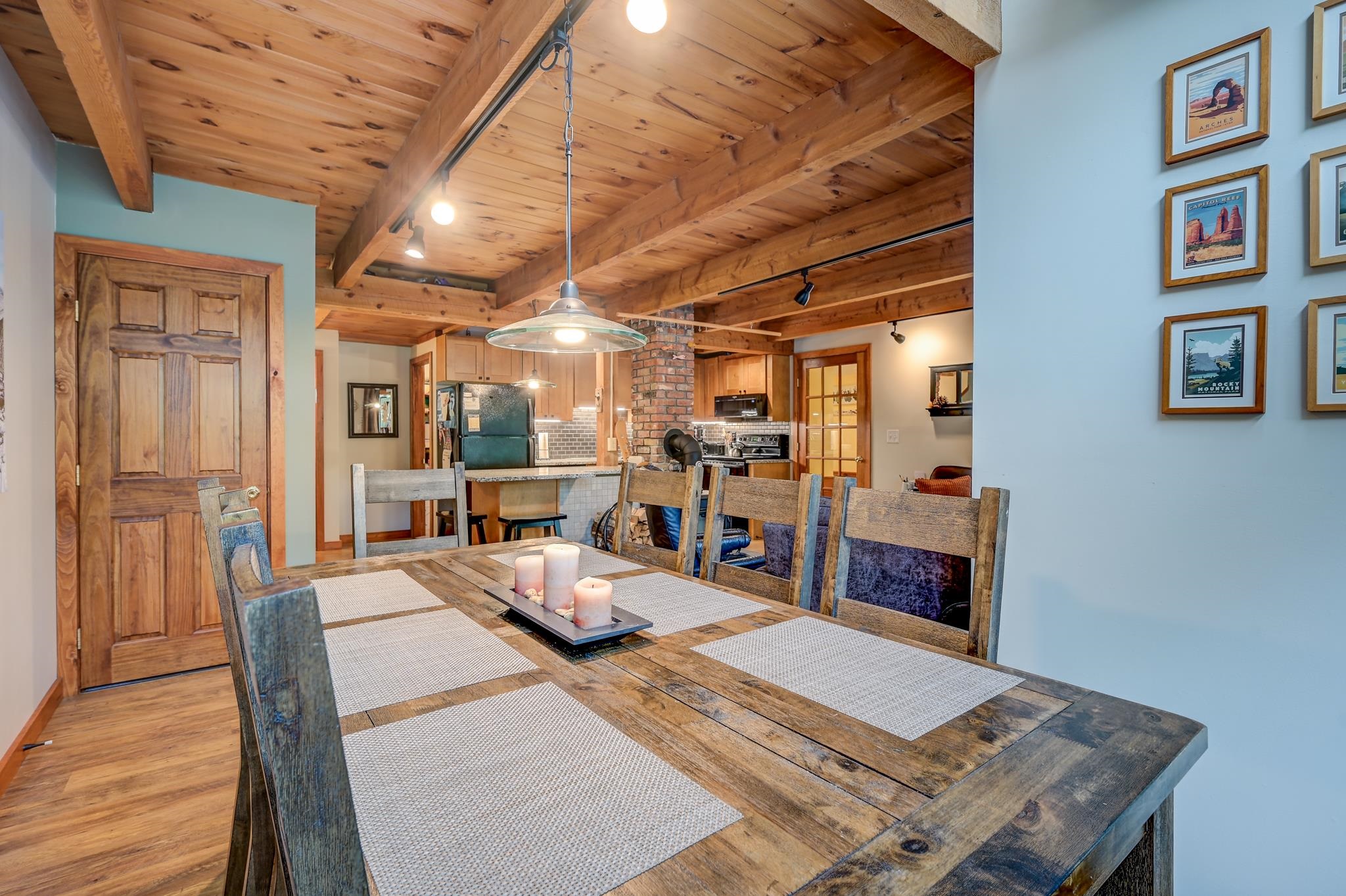 Dining area with a rustic wood table beneath exposed beams, opening to a kitchen with a brick chimney.