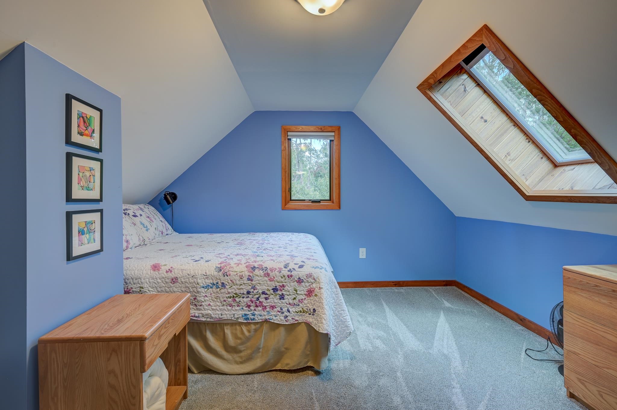 Attic bedroom with blue walls, a floral bedspread, wood furniture, and a large angled skylight.