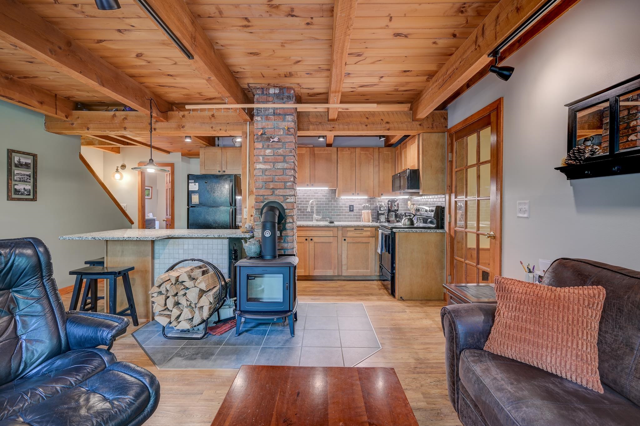 Open-plan living room and kitchen with exposed wood beams, a brick chimney, and a freestanding wood stove.