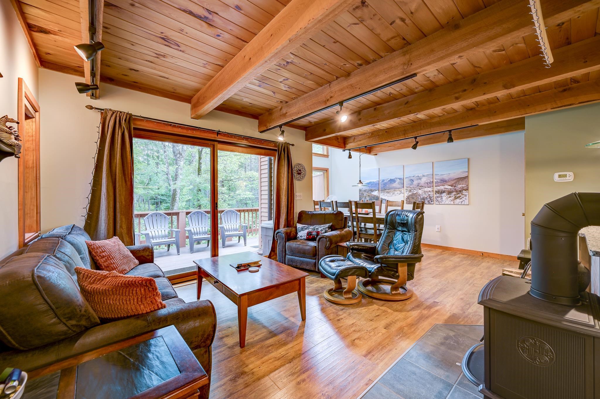 Living room with exposed timber beams, a wood stove, and sliding doors to a wooded deck.