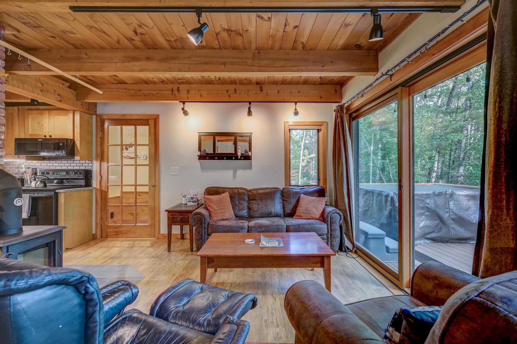 Furnished living room with exposed wood beams, adjoining kitchen, and sliding doors to a wooded deck.