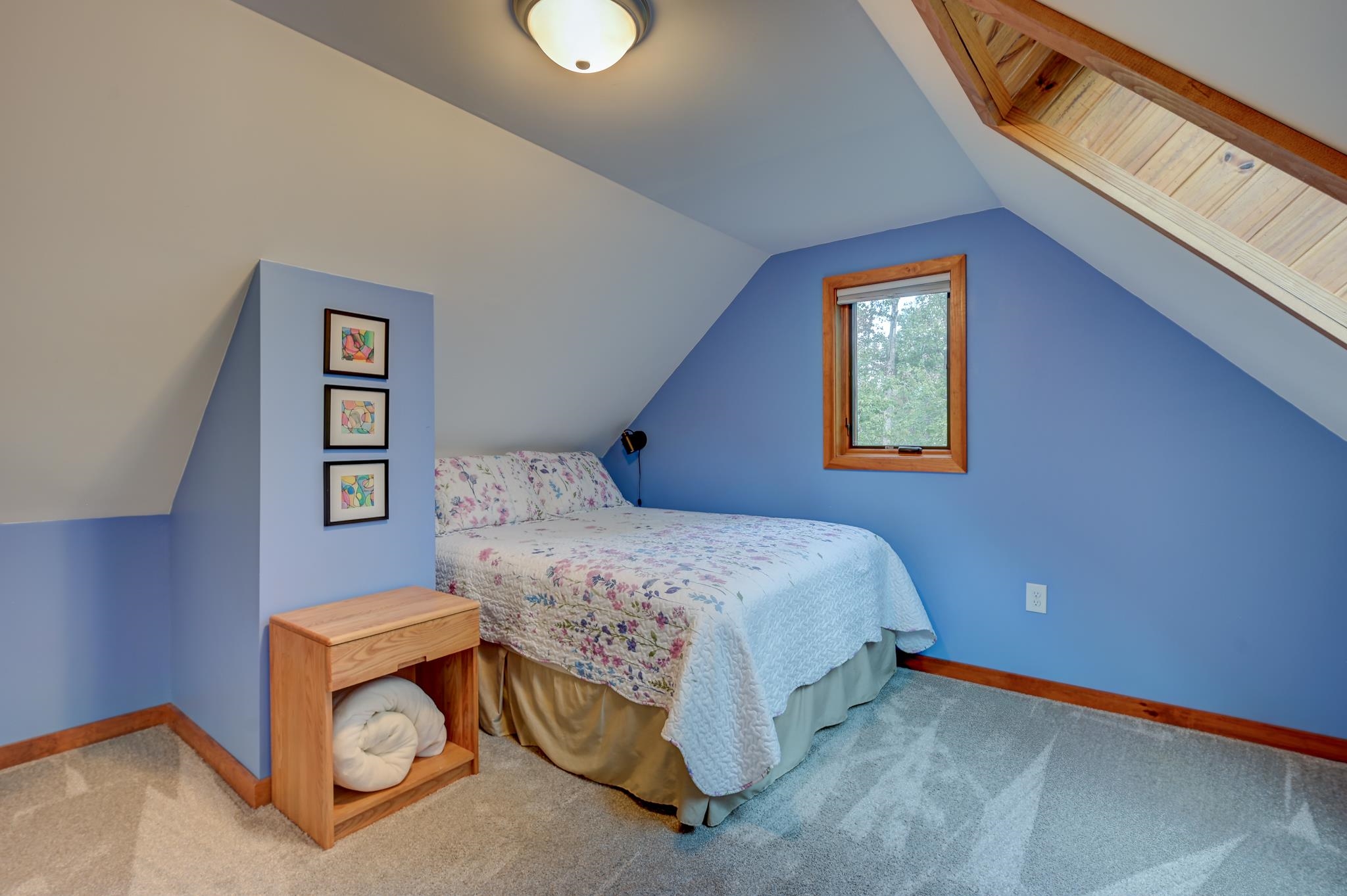 Attic bedroom with blue walls, floral bedspread, sloped ceiling, and wood-trimmed window.