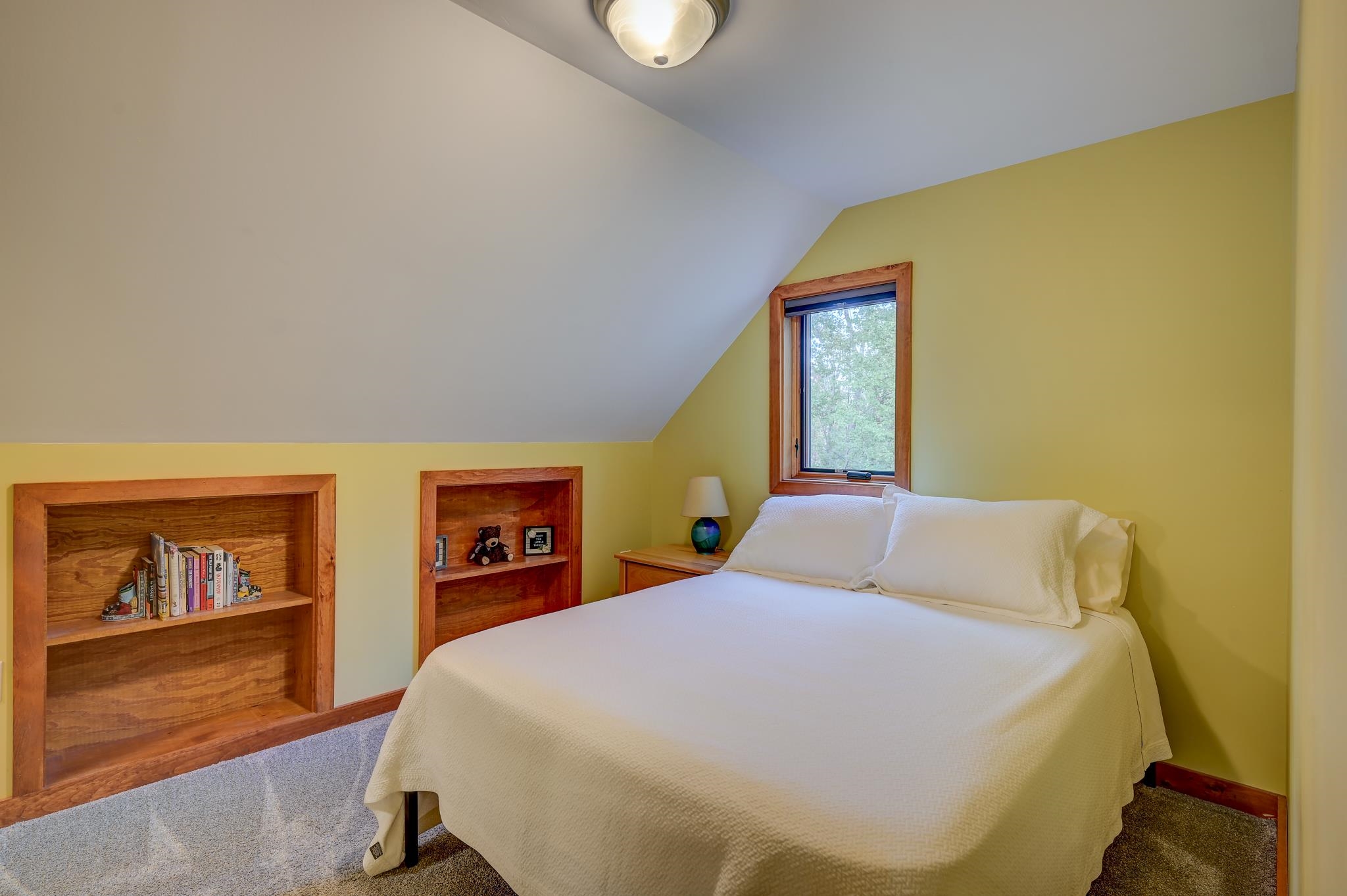 Bedroom with a white bed, sloped ceiling, window, and built-in wooden bookshelves.