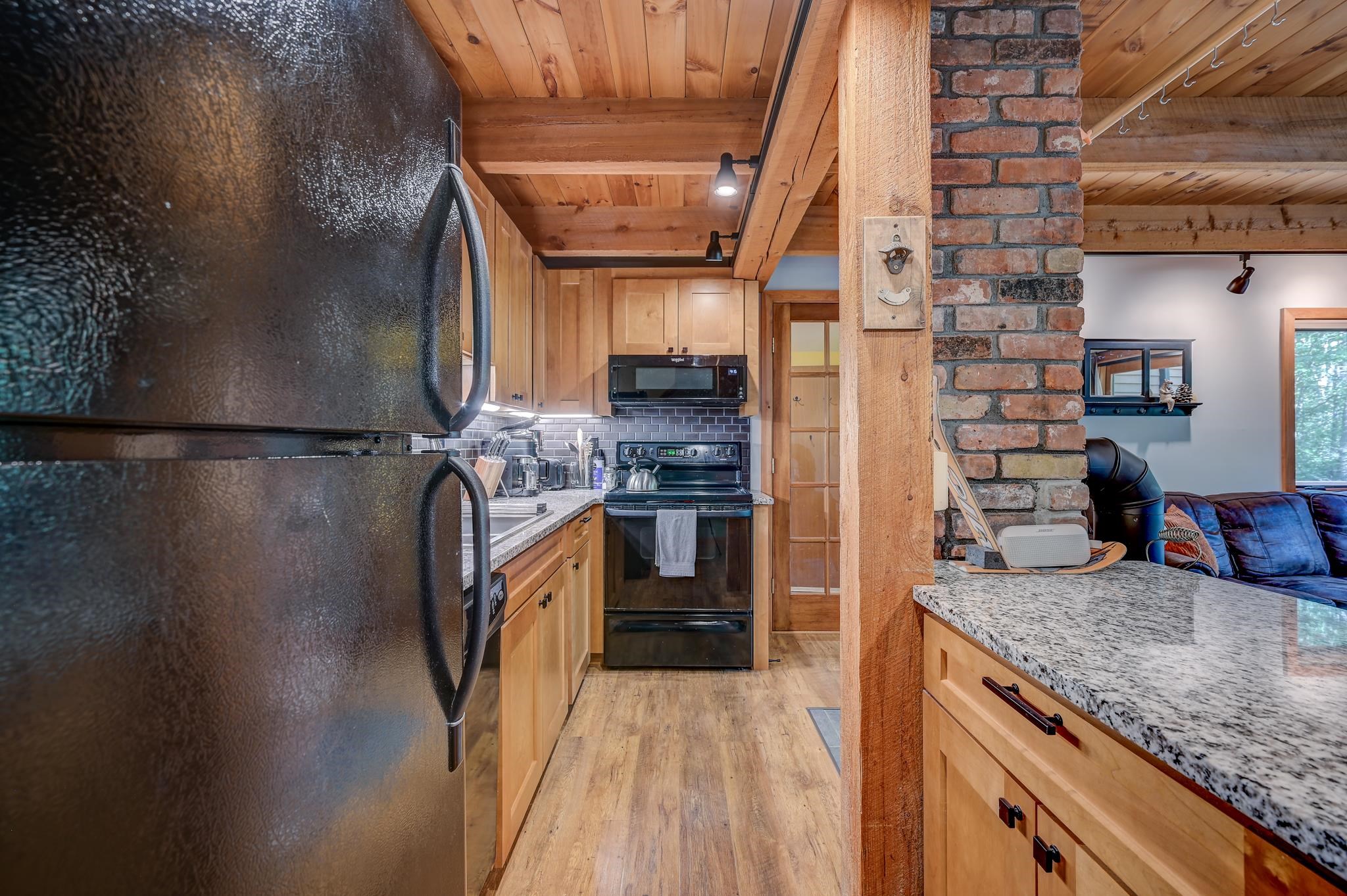 Kitchen with wood cabinetry, granite counters, black appliances, and a brick column beside the living area.