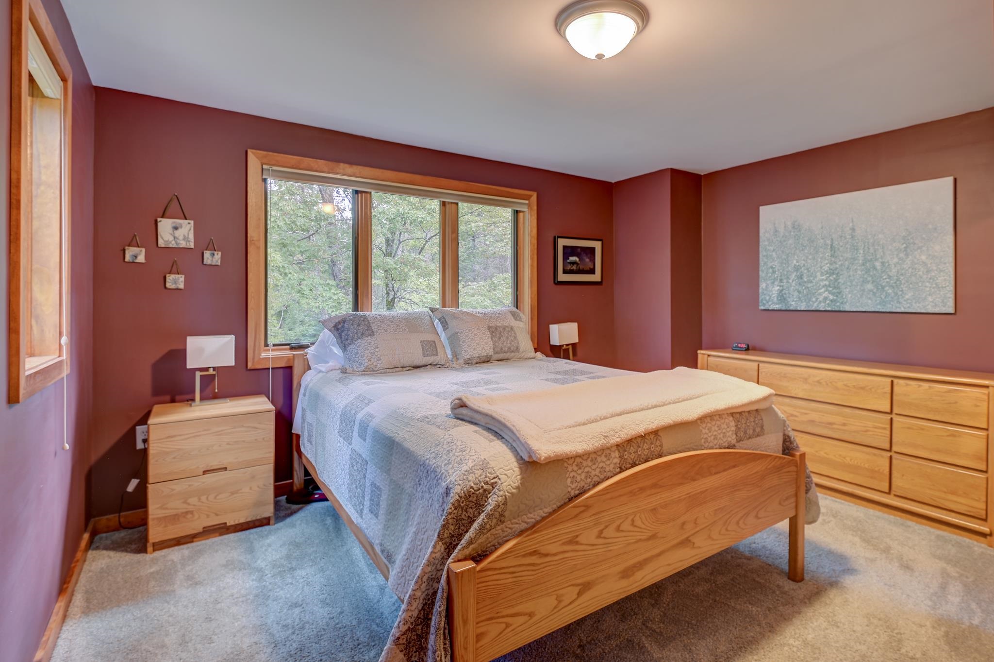 Bedroom with a wooden bed and gray patterned bedding beneath a wide window overlooking trees.