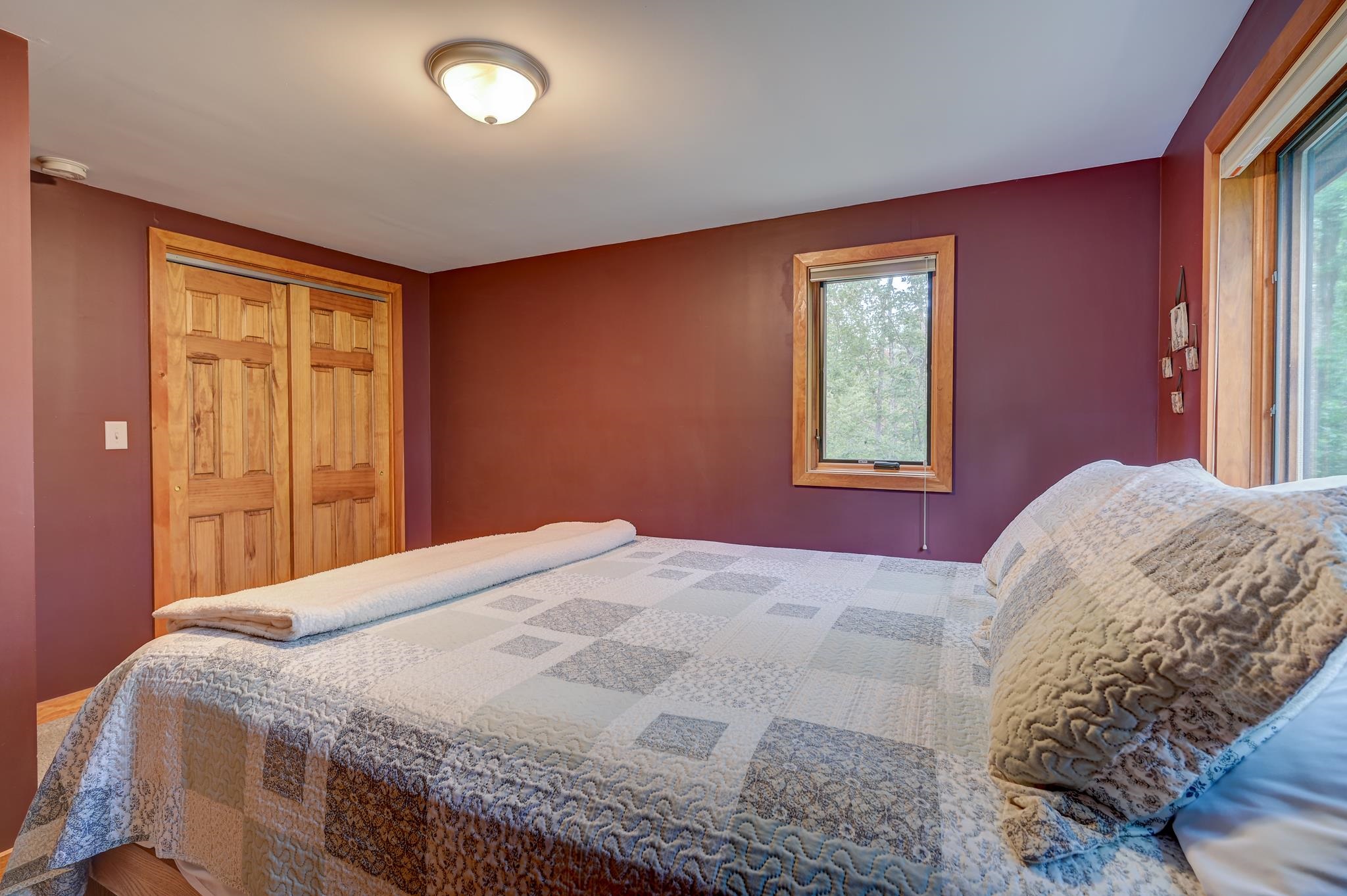Bedroom with a patterned quilted bed, burgundy walls, wood closet doors, and windows overlooking trees.