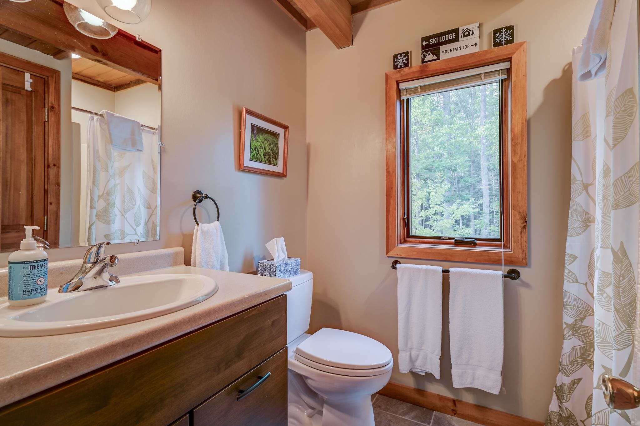 Bathroom with a wood vanity, patterned shower curtain, and window overlooking leafy trees.