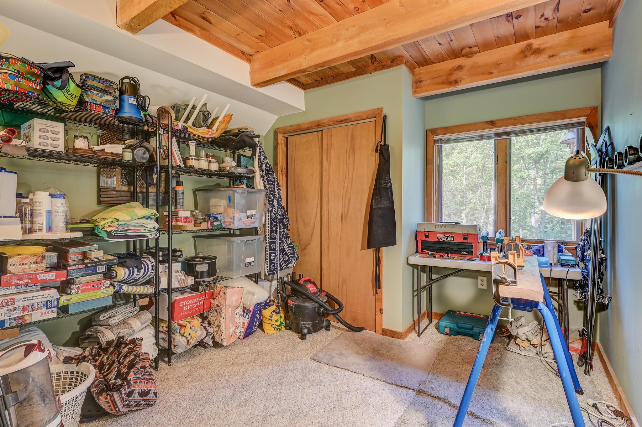 Craft room with exposed wood beams, packed storage shelves, and a worktable beside a large window.