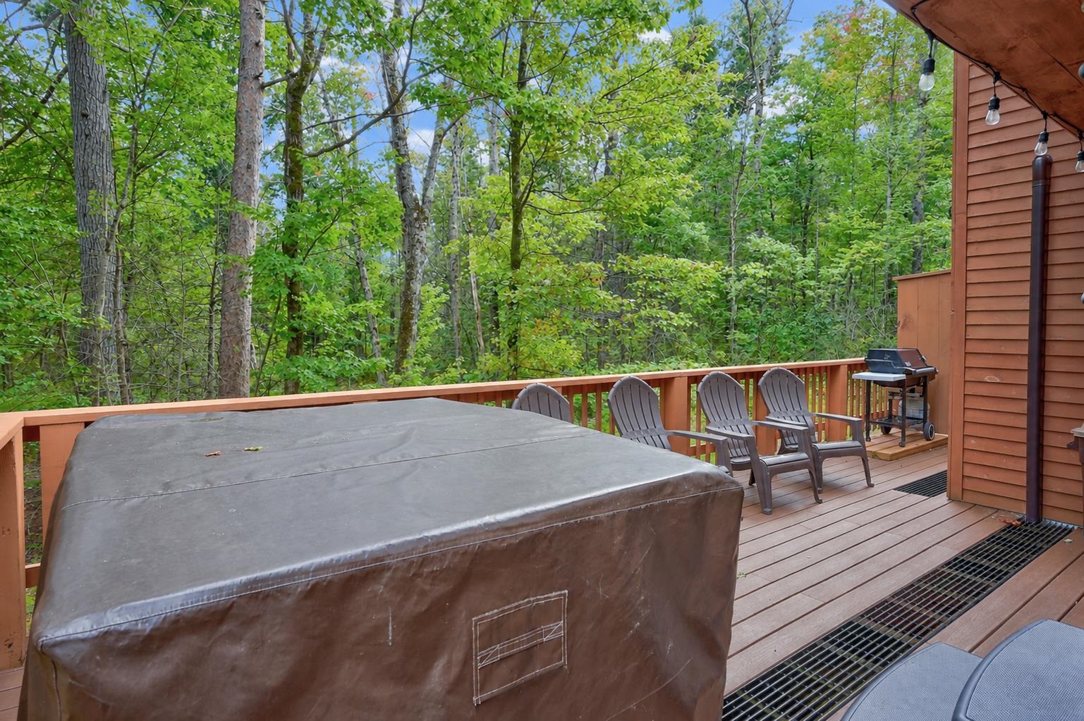 Wooden deck with a covered hot tub, Adirondack chairs, and grill overlooking leafy woods.