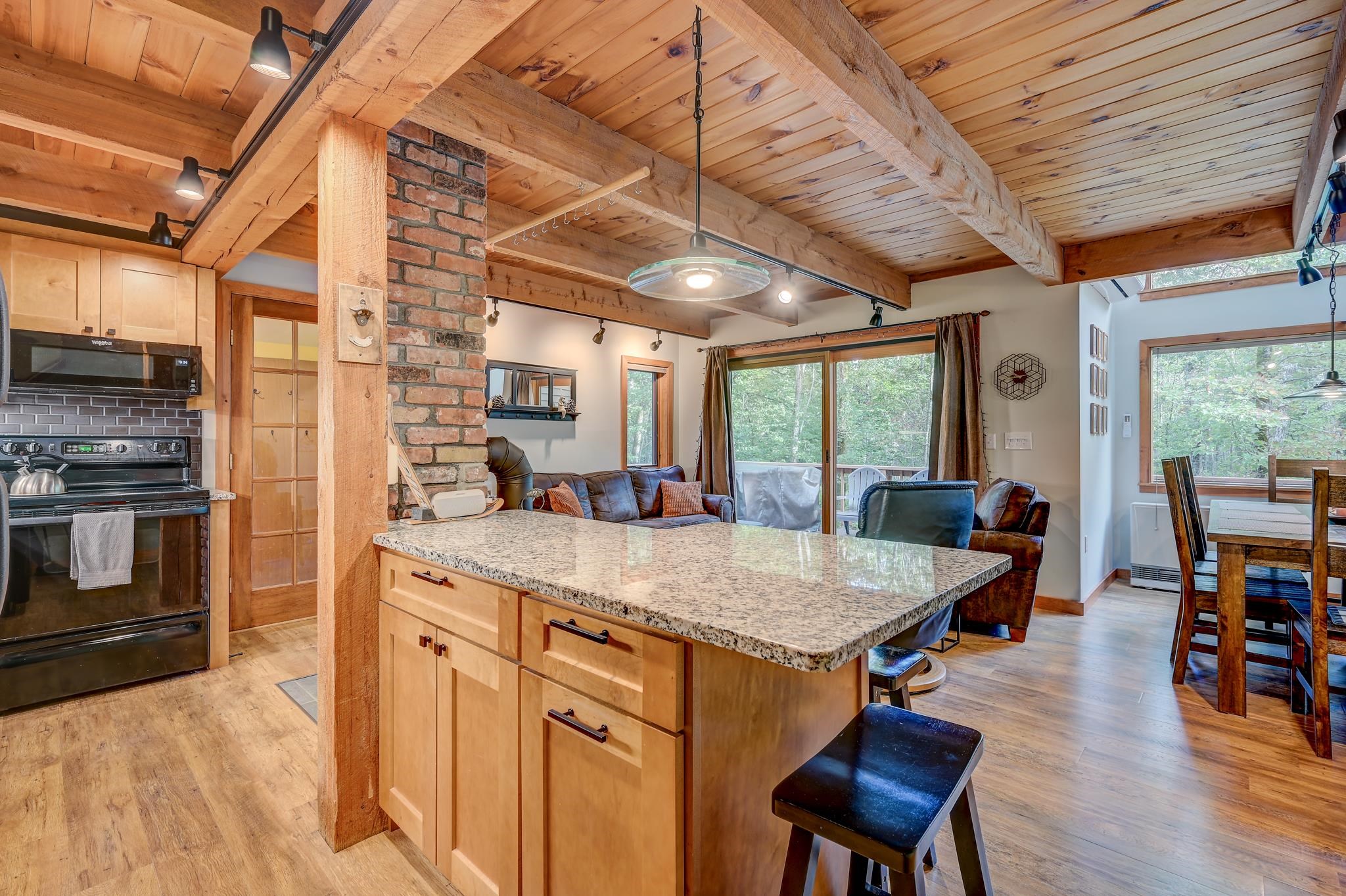 Open-plan kitchen and living area with a granite island, exposed timber beams, and wooded views.