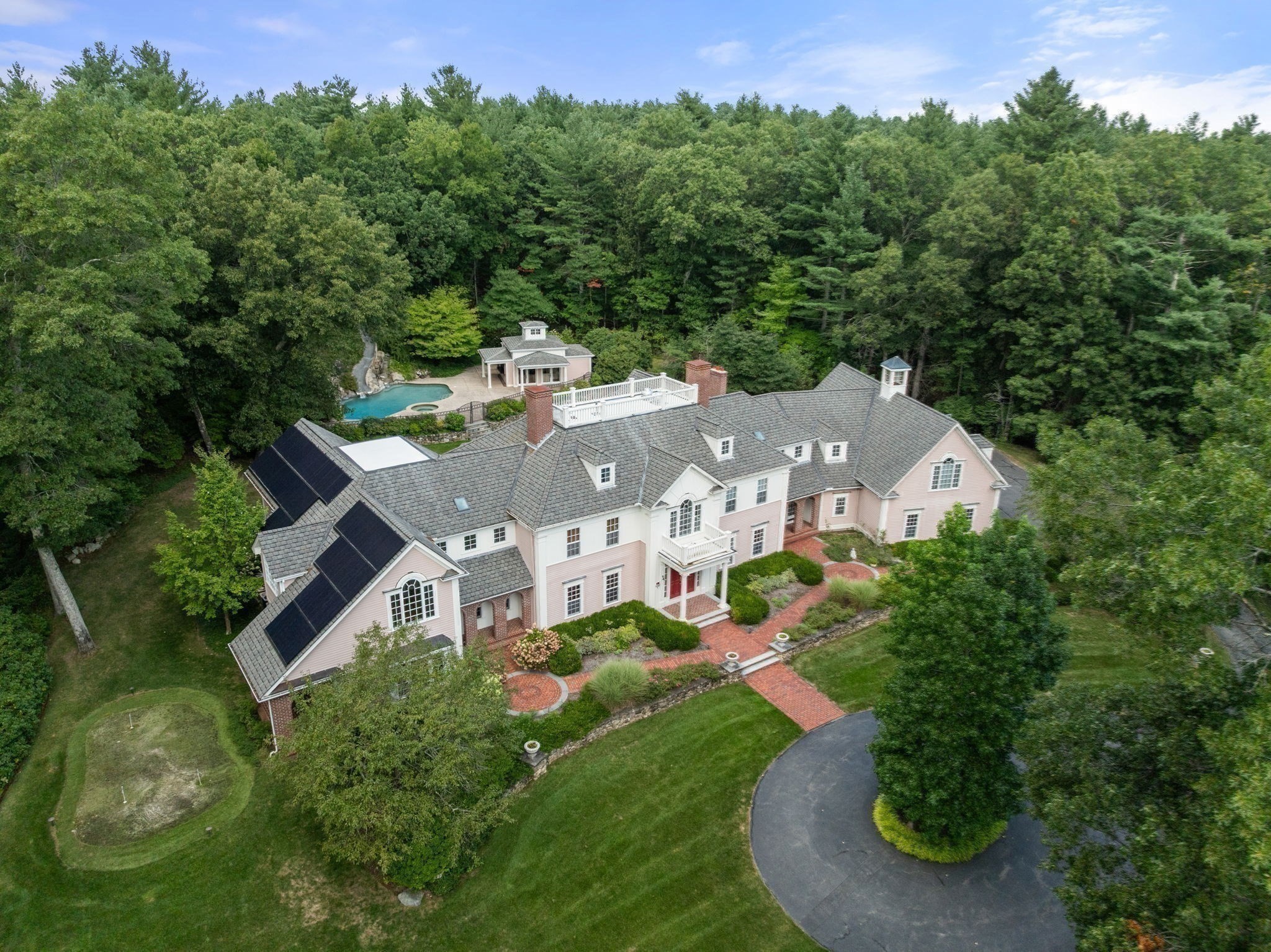 Aerial view of a large pink-and-white home nestled in woods, with a swimming pool and curved driveway.