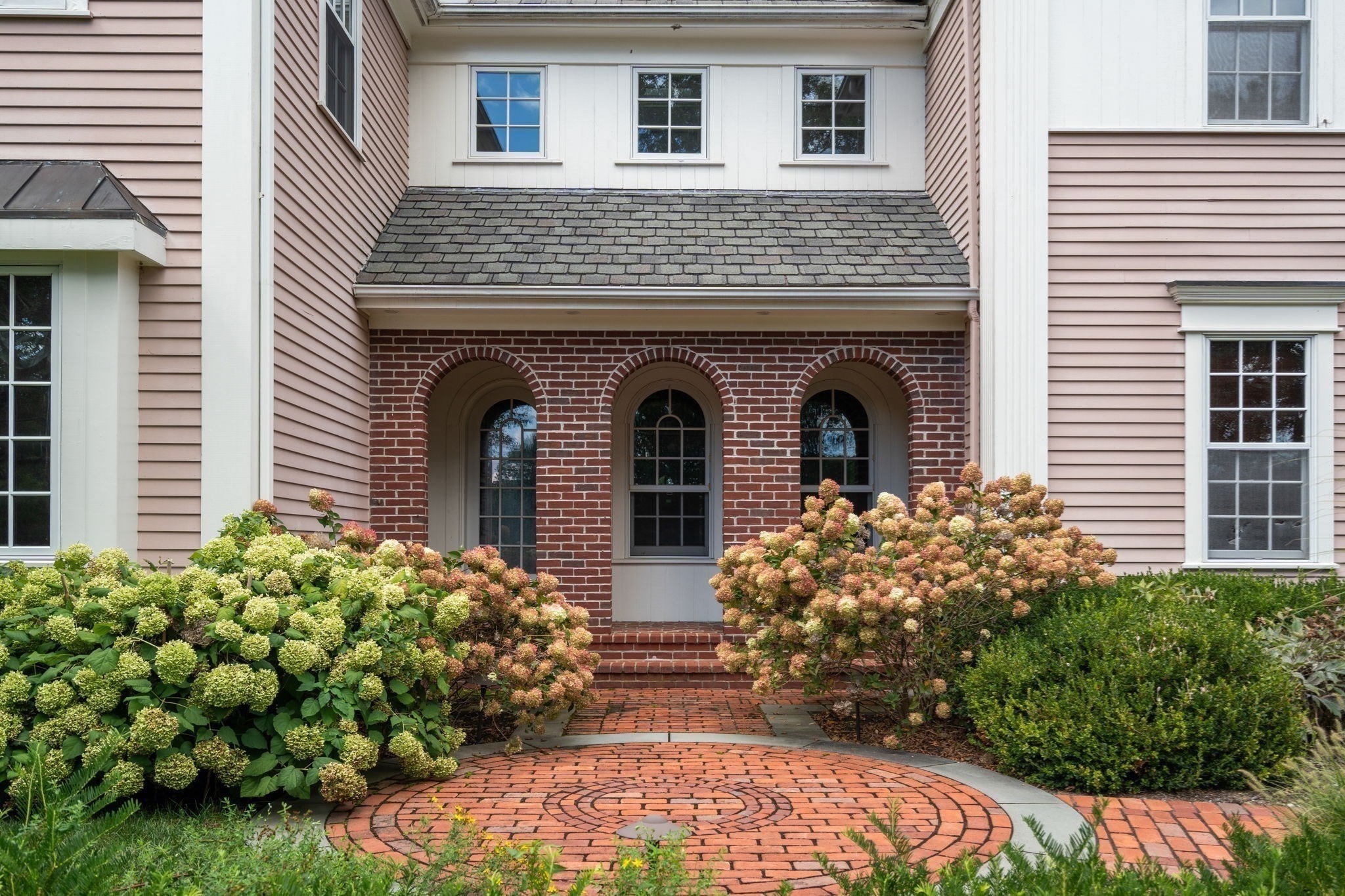 Home entrance with three arched windows, brick steps, a circular brick walkway, and flowering shrubs.