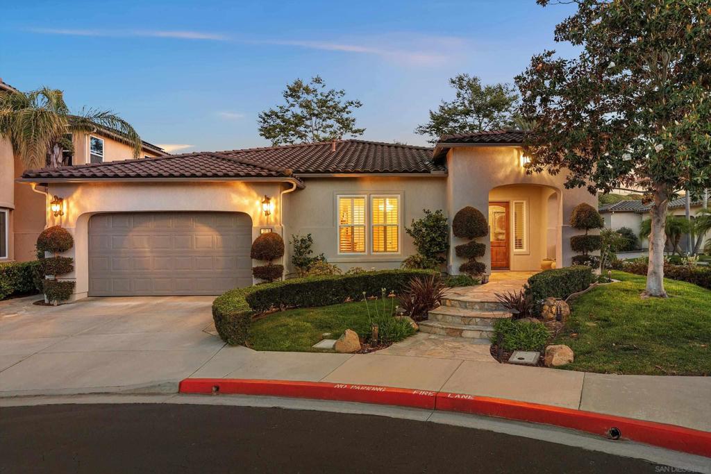 Stucco home with a tiled roof, double garage, and warmly lit landscaped entry at dusk.