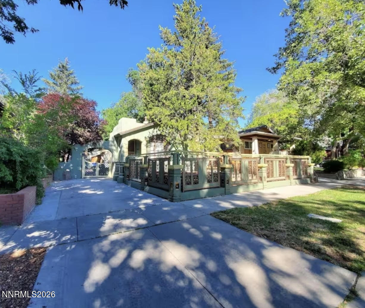 House with a broad raised porch and decorative railing, shaded by mature trees beside a wide concrete driveway.