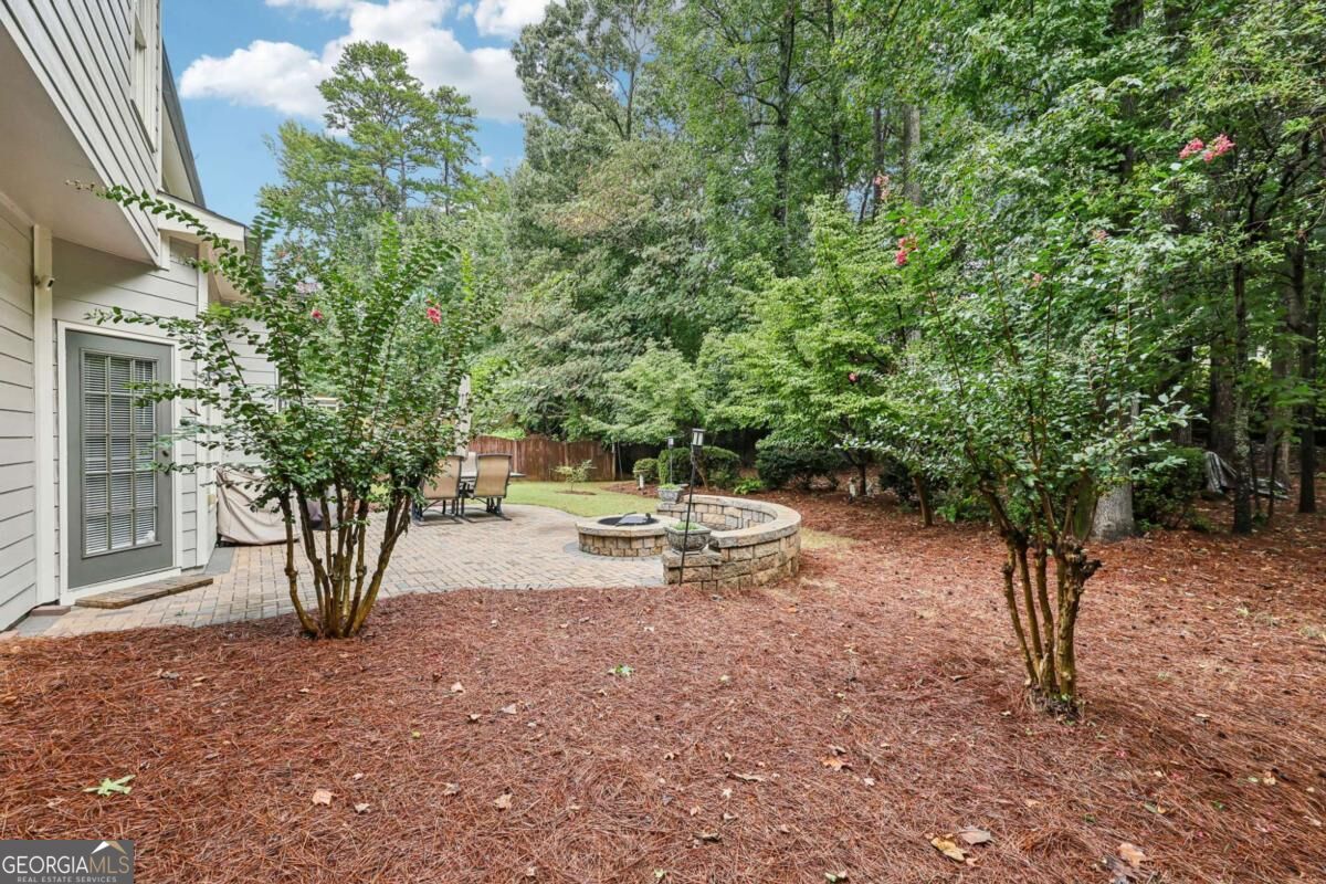 Backyard patio with a circular stone fire pit and curved seating wall, surrounded by mature trees.