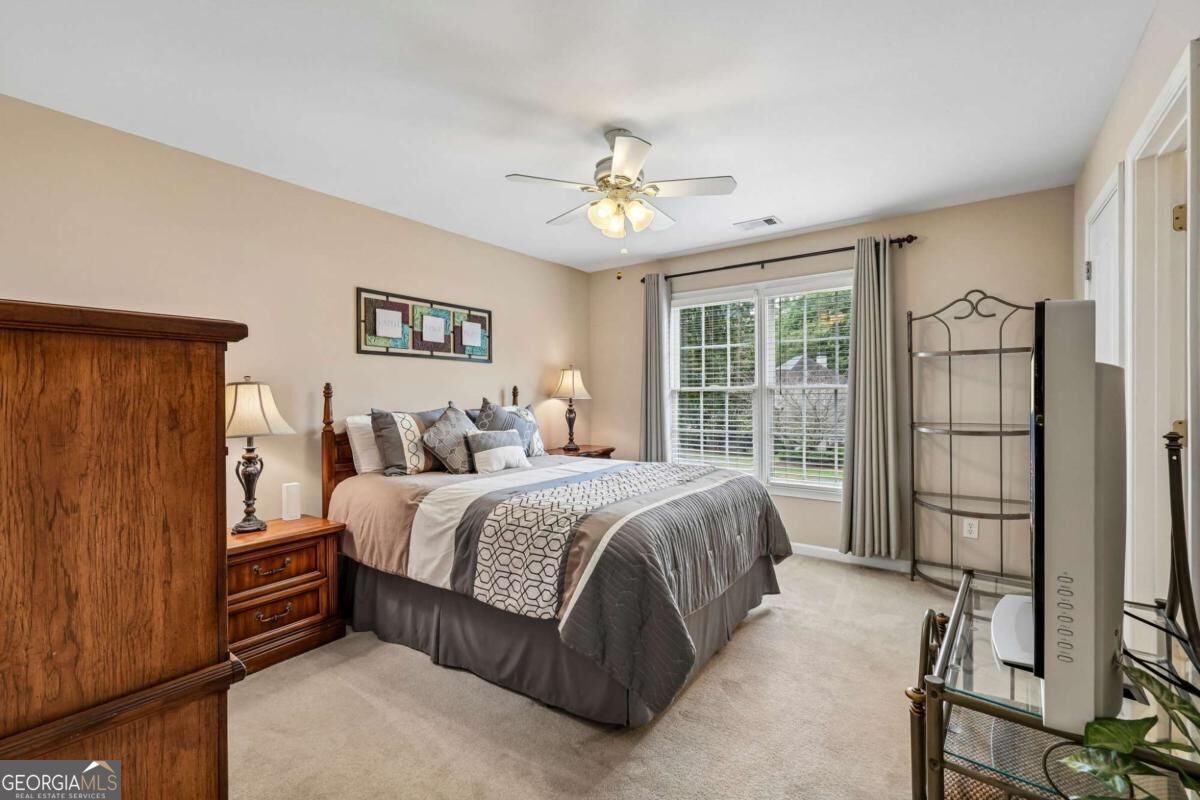 Furnished bedroom with gray and white bedding, wood furniture, and a wide window overlooking greenery.