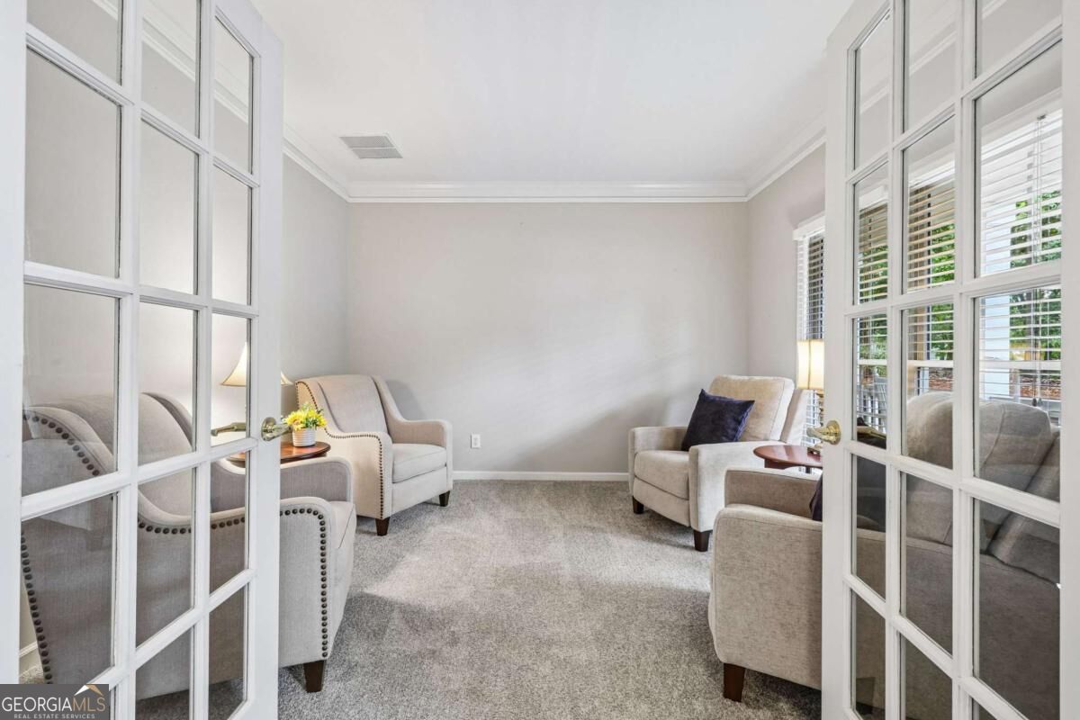 Staged sitting room with pale armchairs, gray carpet, and tall glass-paneled French doors.