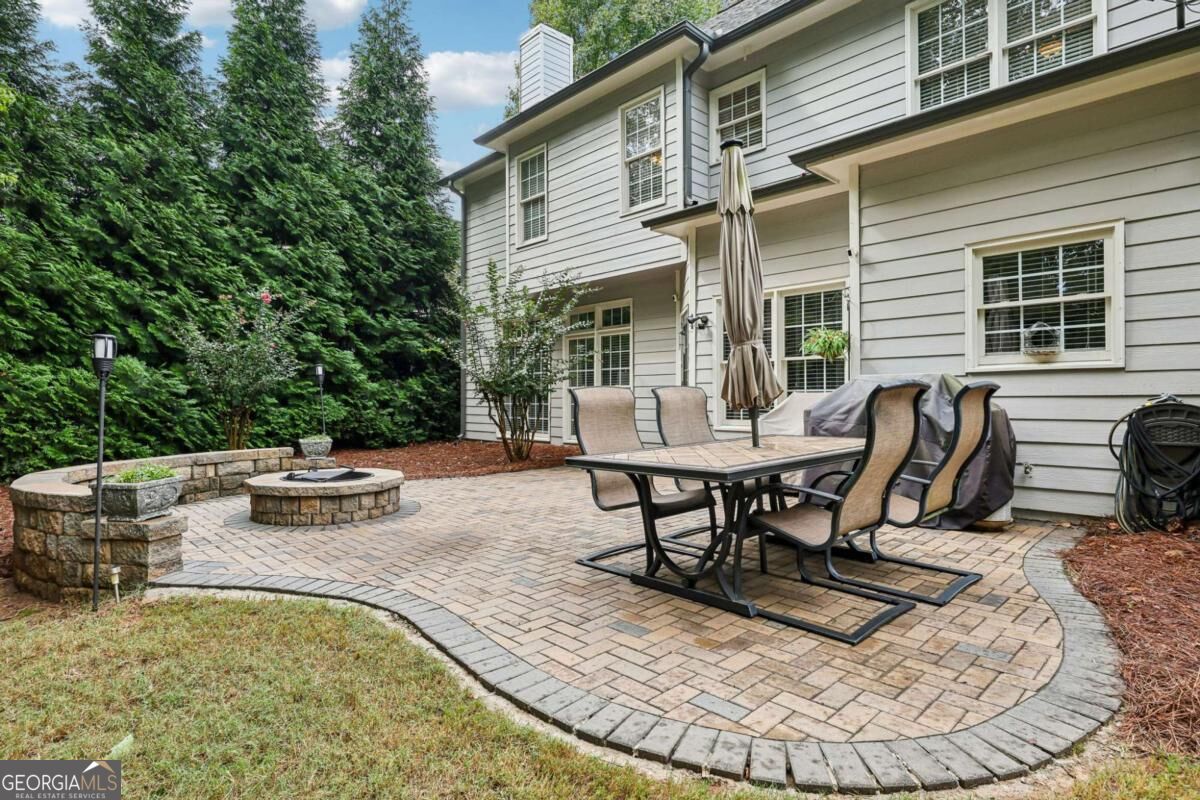 Backyard brick patio with outdoor dining set and circular stone fire pit beside a two-story house.