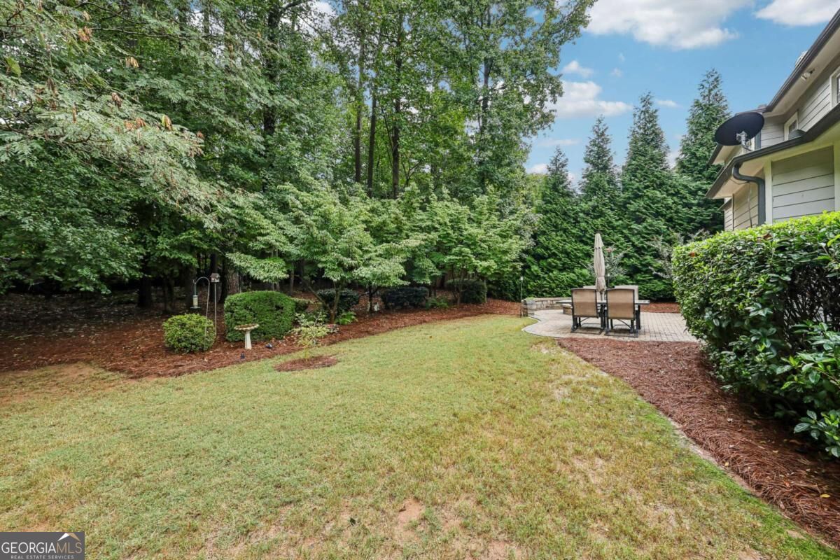Backyard lawn bordered by mature trees, with a paved patio and outdoor dining set.