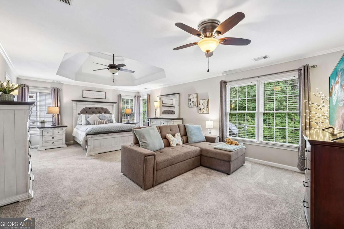 Staged bedroom with a gray bed, taupe sectional, two ceiling fans, and windows overlooking greenery.