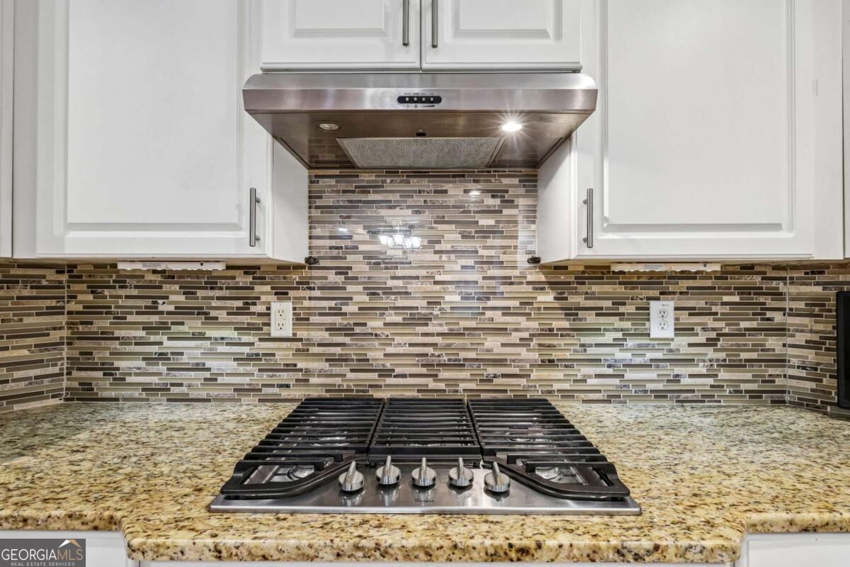 Kitchen cooktop beneath a stainless steel range hood, with granite counters and a mosaic tile backsplash.