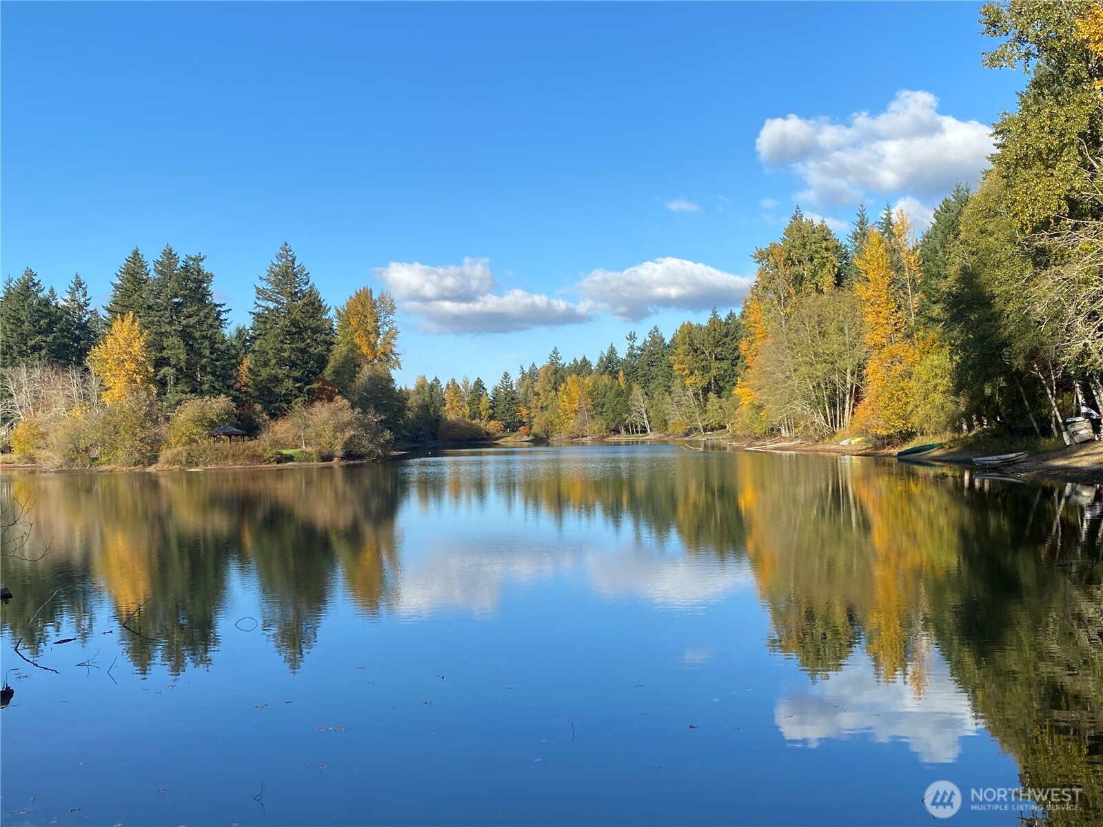 Wooded pond reflecting autumn trees beneath a bright blue sky.