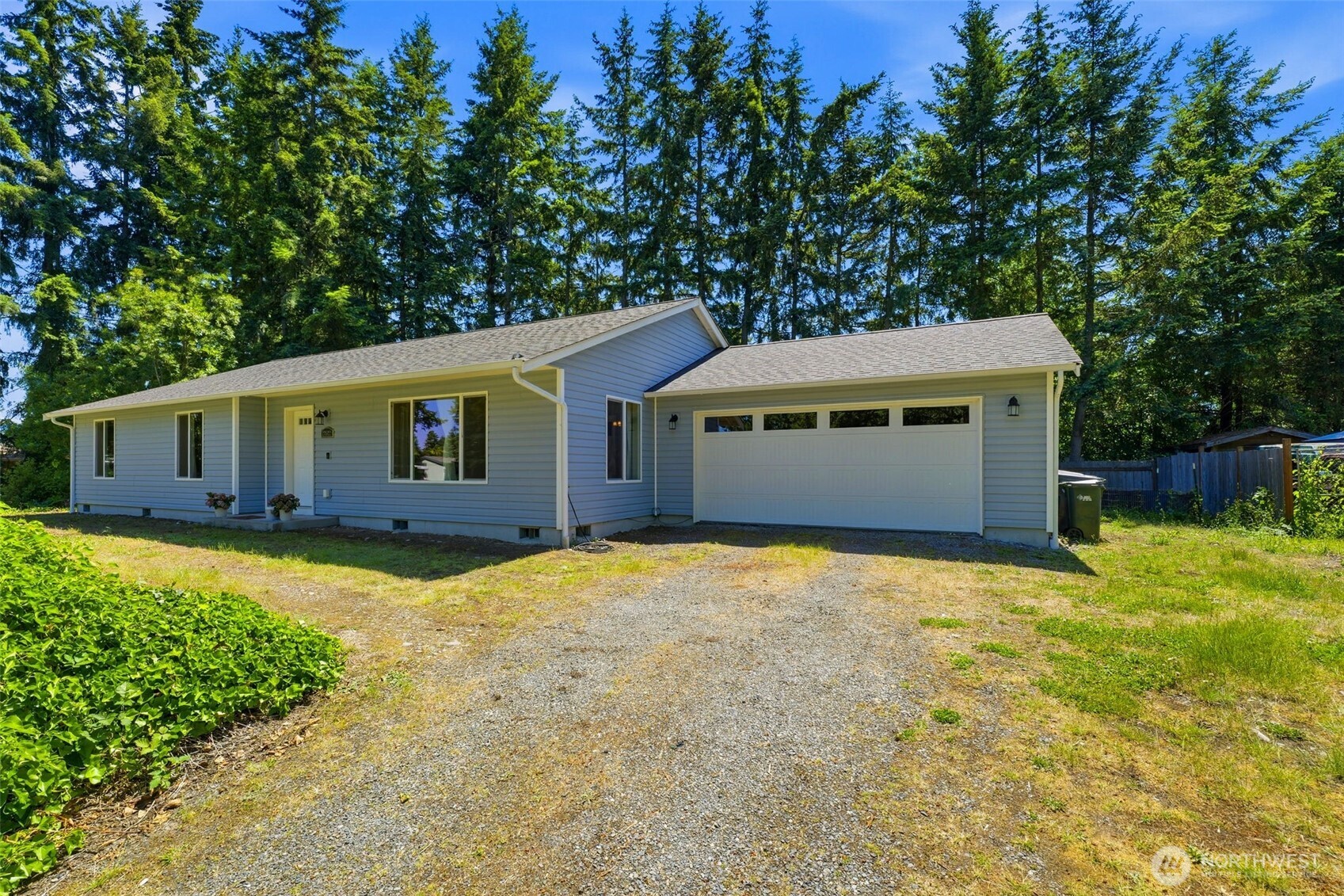 Single-story blue-gray house with an attached garage and gravel driveway, backed by tall evergreens.