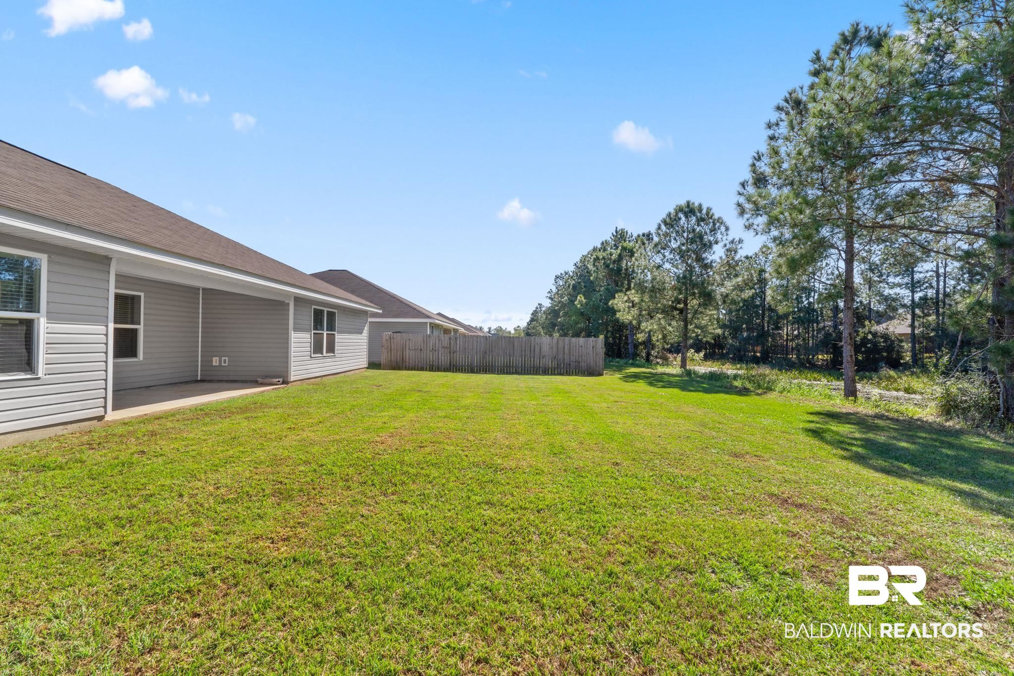 Grassy backyard beside a gray-sided house, with a covered patio, privacy fence, and pine trees.