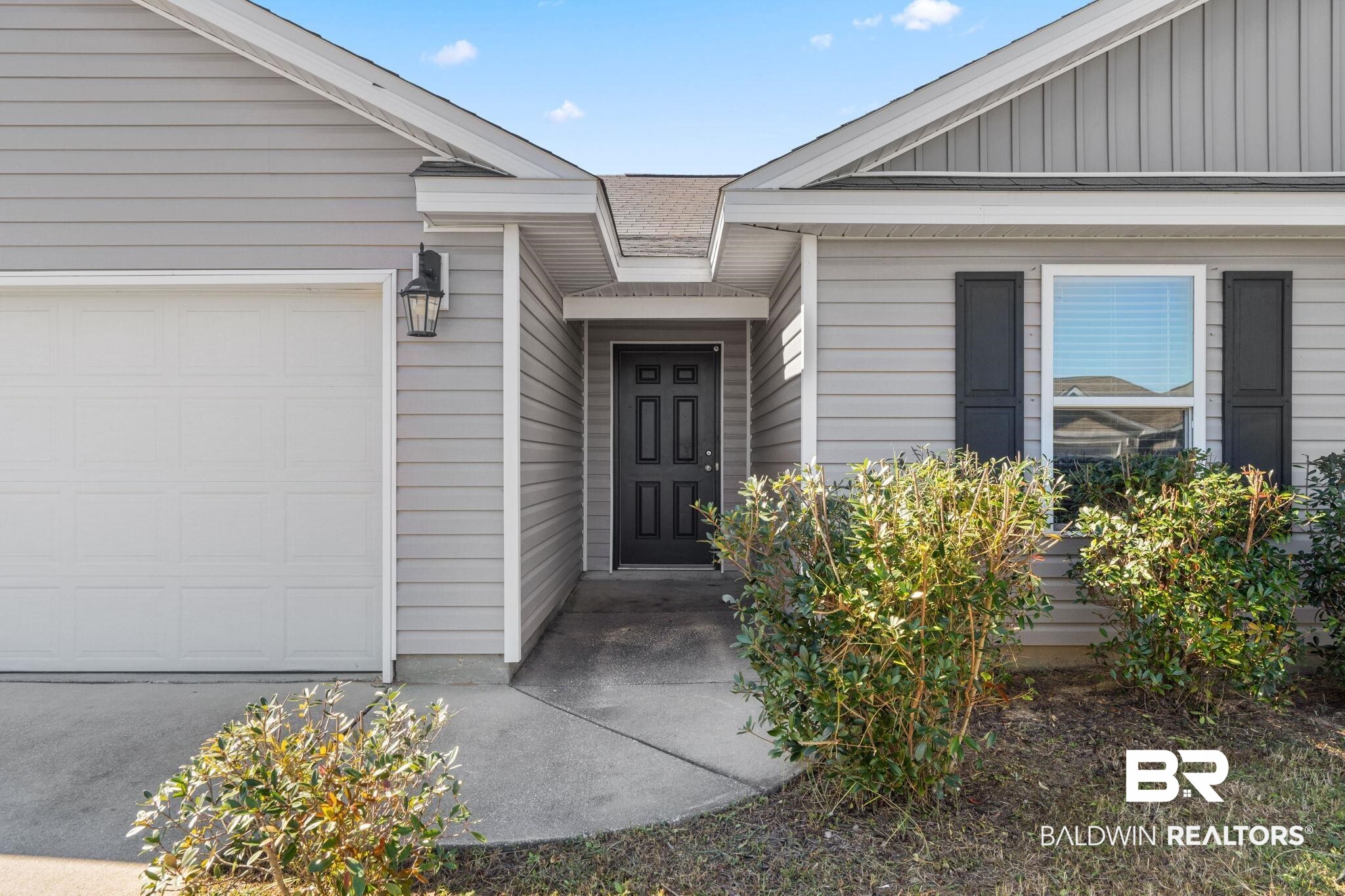 Gray home exterior with a white garage door, covered entry, black front door, and foundation shrubs.