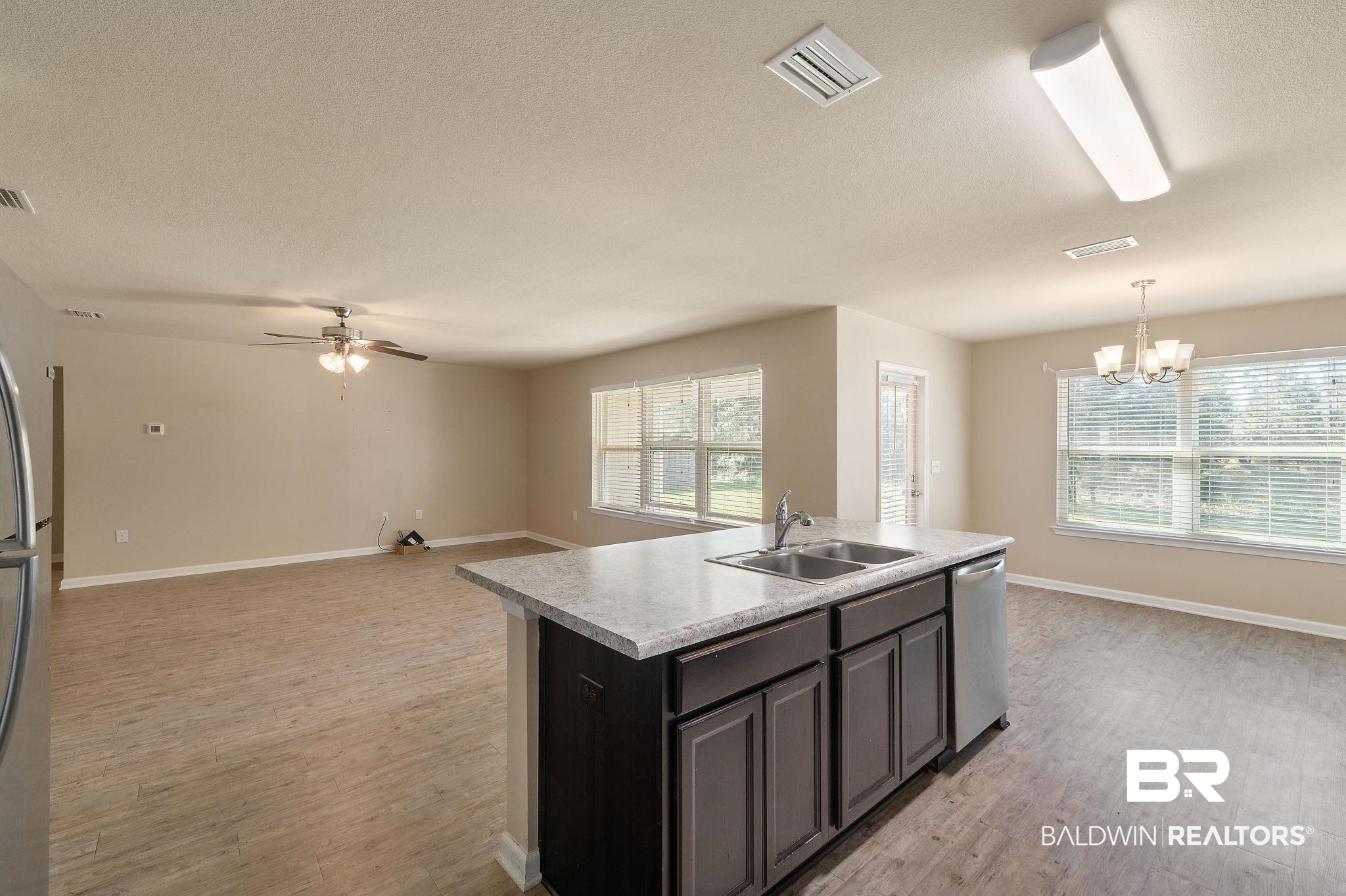 Vacant kitchen with a dark island, double-basin sink, wood-look floors, and large windows.