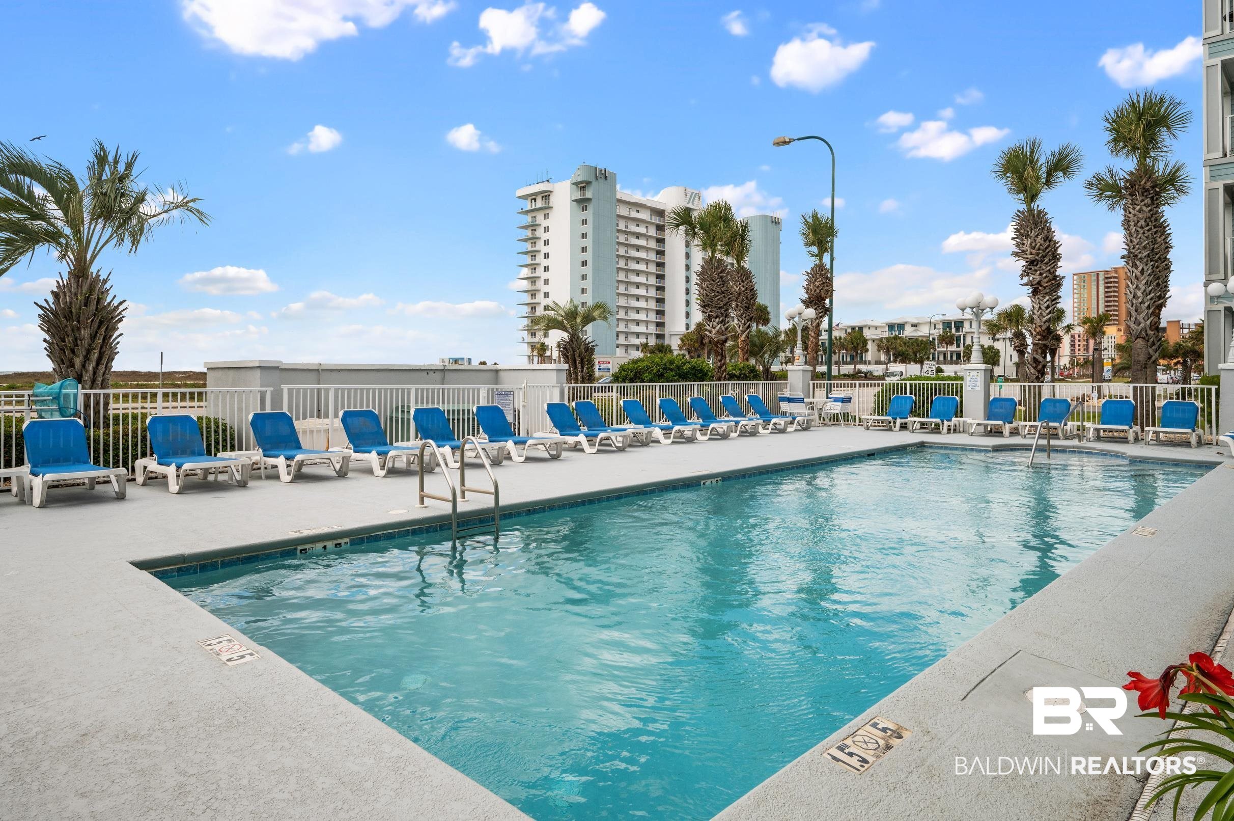 Outdoor pool lined with blue lounge chairs, with palm trees and condominium towers behind it.
